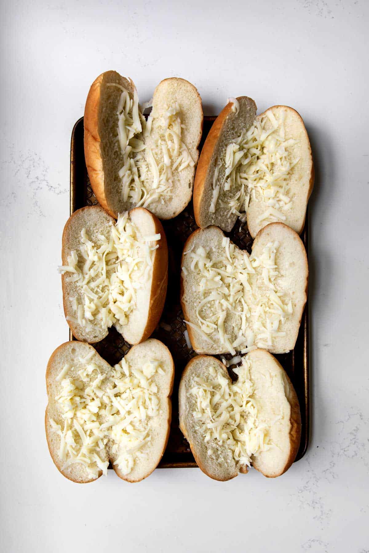 Six sausage buns with grated provolone cheese on a baking sheet.