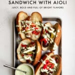 4 Italian Sausage Sandwiches with roasted peppers and onions with basil mayo on top, along with a small bowl of basil aioli sauce on a baking sheet with text description.