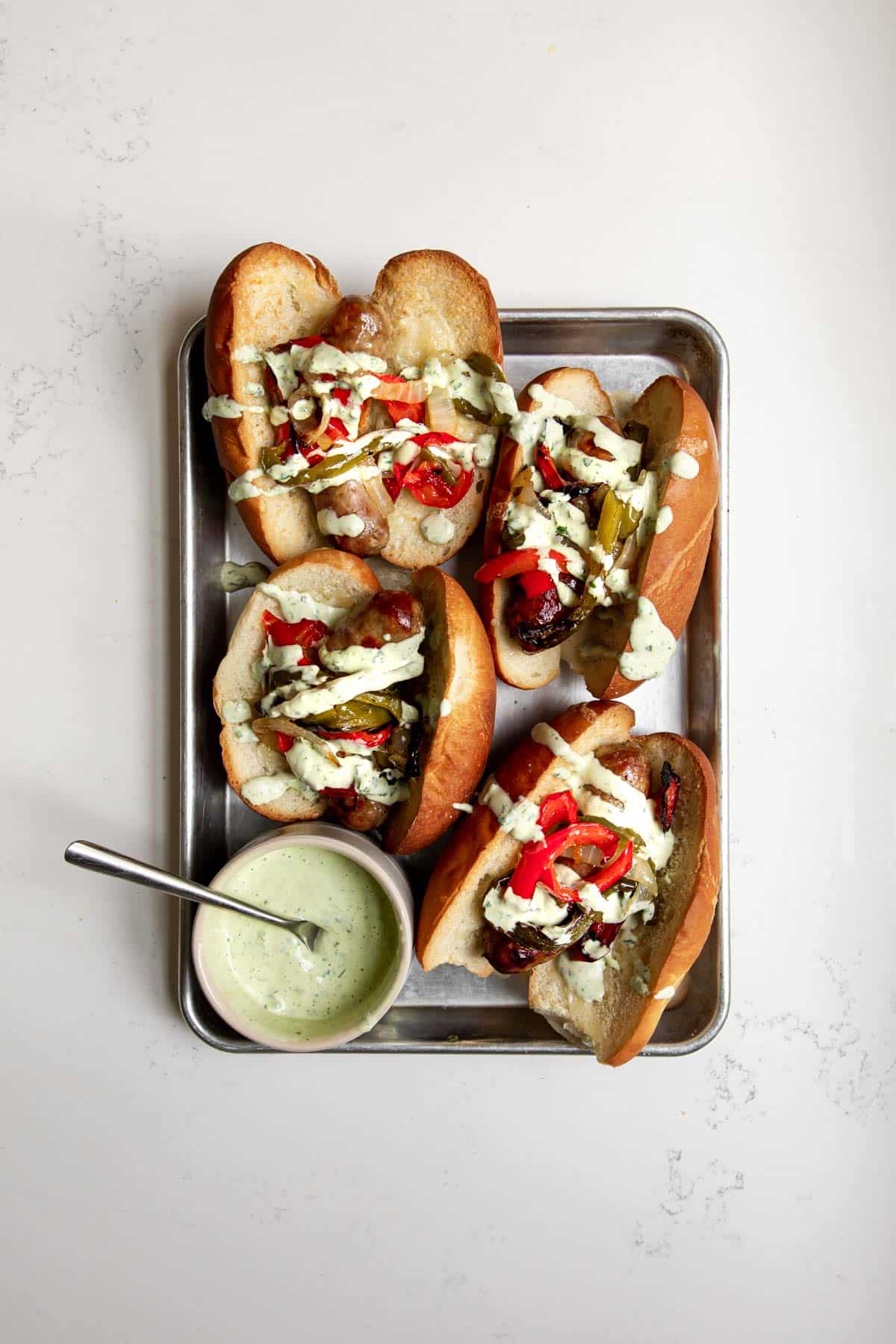 4 Italian Sausage Sandwiches with roasted peppers and onions with basil mayo on top, along with a small bowl of basil aioli sauce on a baking sheet.