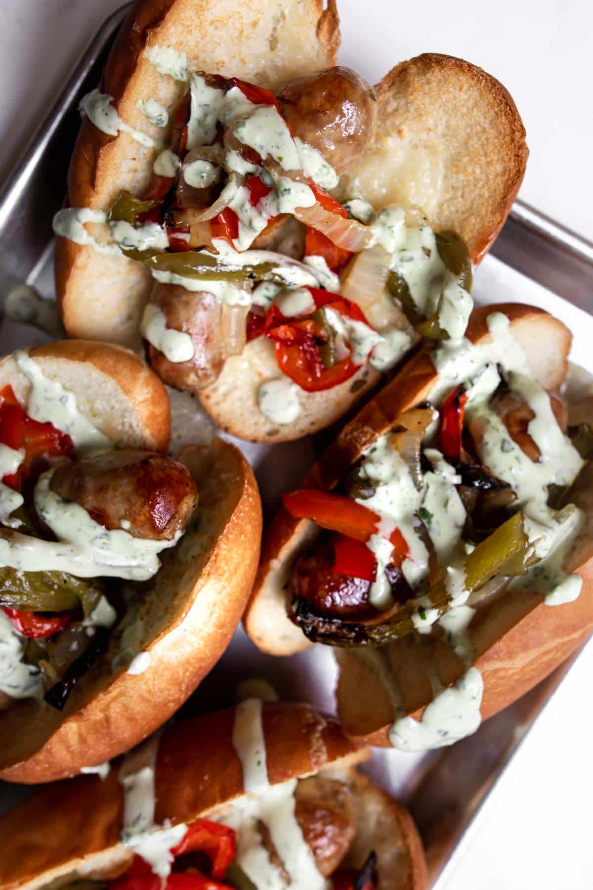 Close up of baked sausages, peppers and onions on a bun with basil mayonnaise sauce on top.