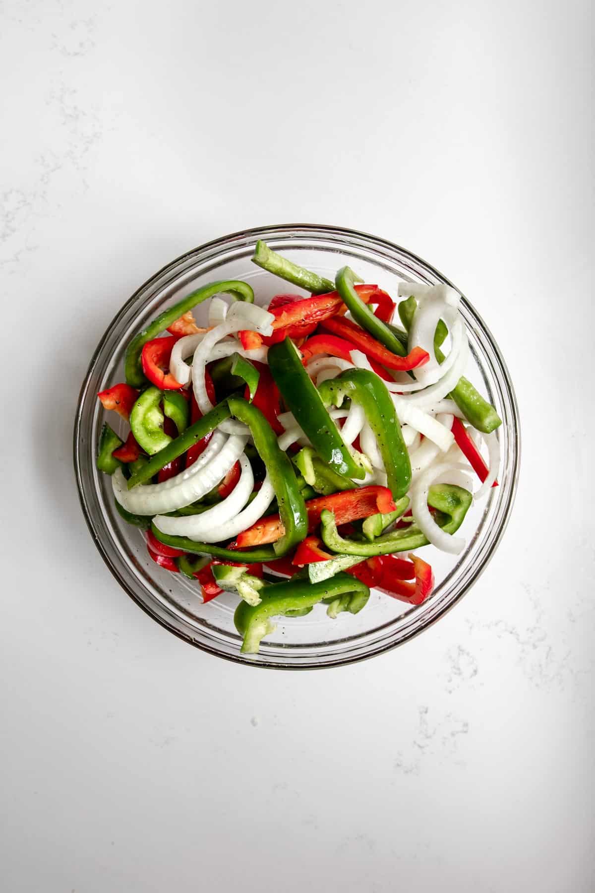 Sliced green peppers, red peppers, and onions, tossed with oil, salt, and pepper, in a glass bowl on white marble background.
