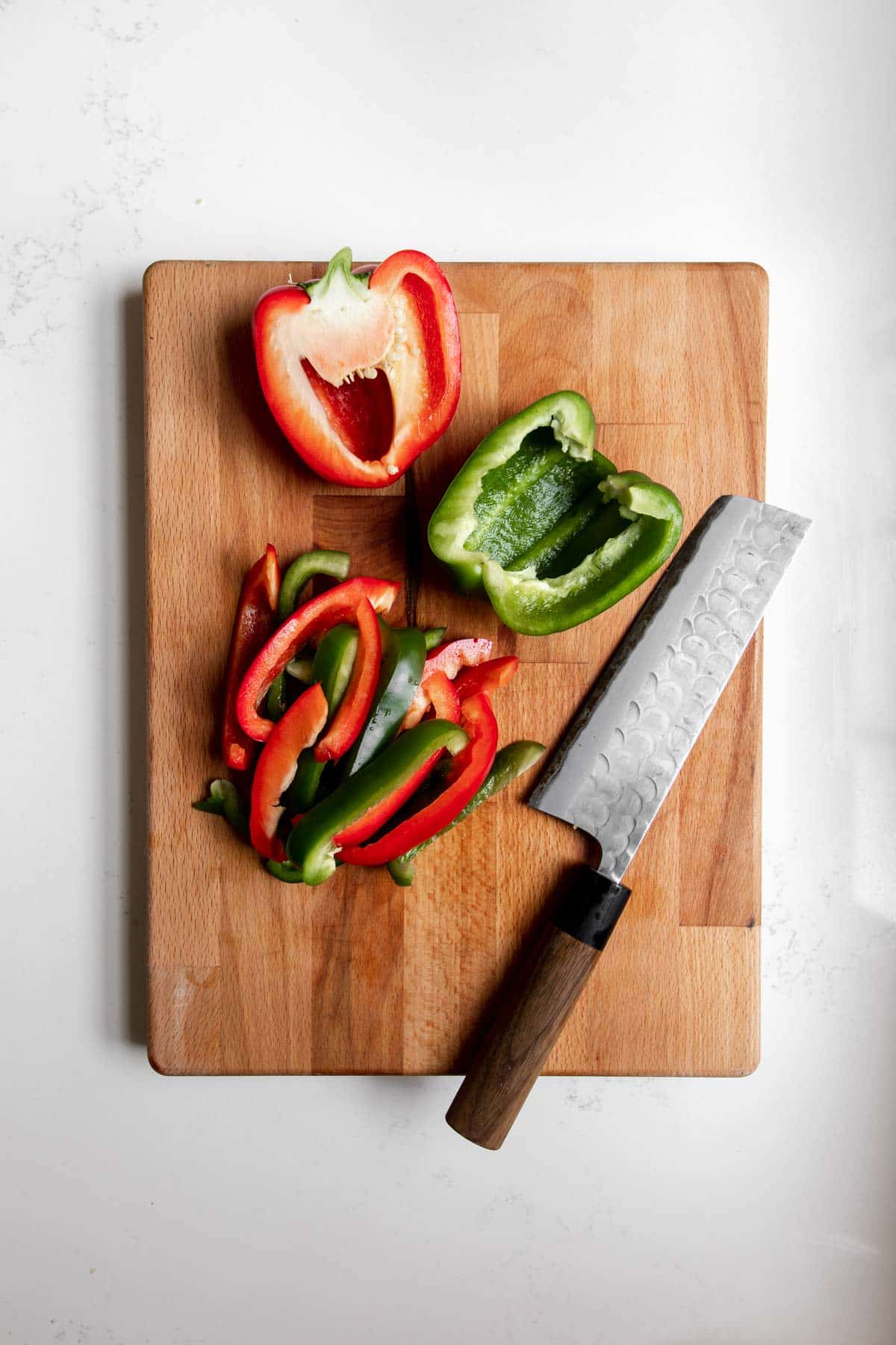 Half a red pepper, green pepper, red and green pepper slices and and Japanese knife on a wooden cutting board.