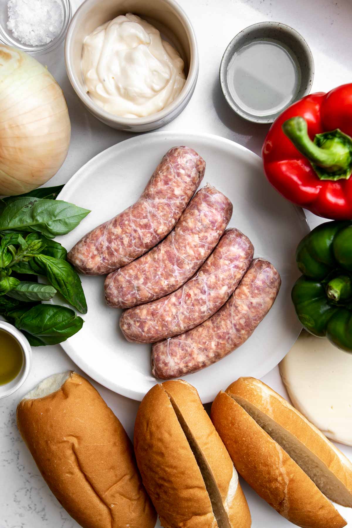 Small plate with 4 sausages, surrounded by 3 sausage buns, olive oil, fresh basil, garlic, onion, sea salt flakes, mayonnaise, pickle juice, red pepper, green pepper, and provolone cheese.