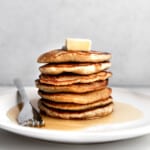 Stack of oat flour pancakes with butter and maple syrup on a white plate with silver fork.