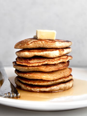 Stack of oat flour pancakes with butter and maple syrup on a white plate with silver fork.