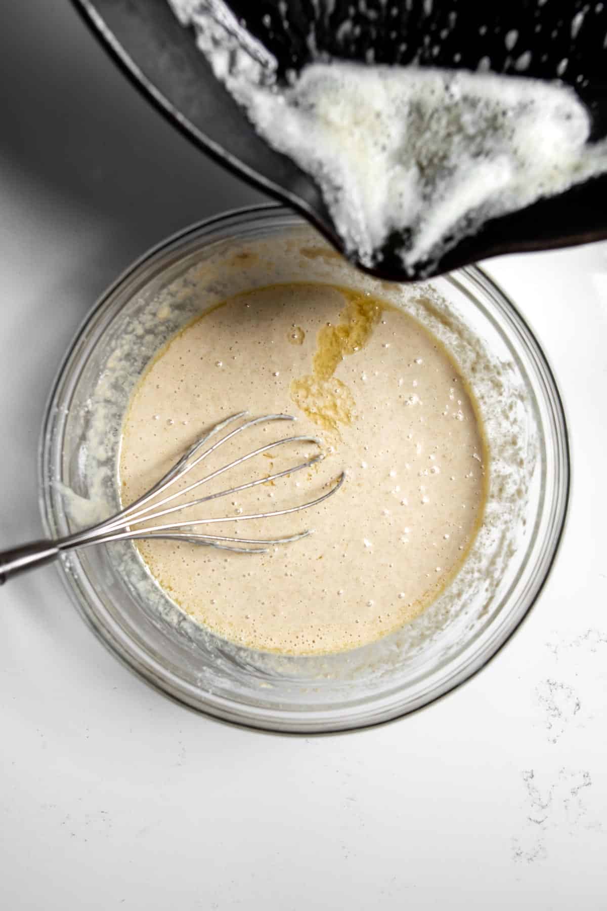 Melted butter being poured in a bowl with a whisk sitting in oat flour pancake batter from a skillet.
