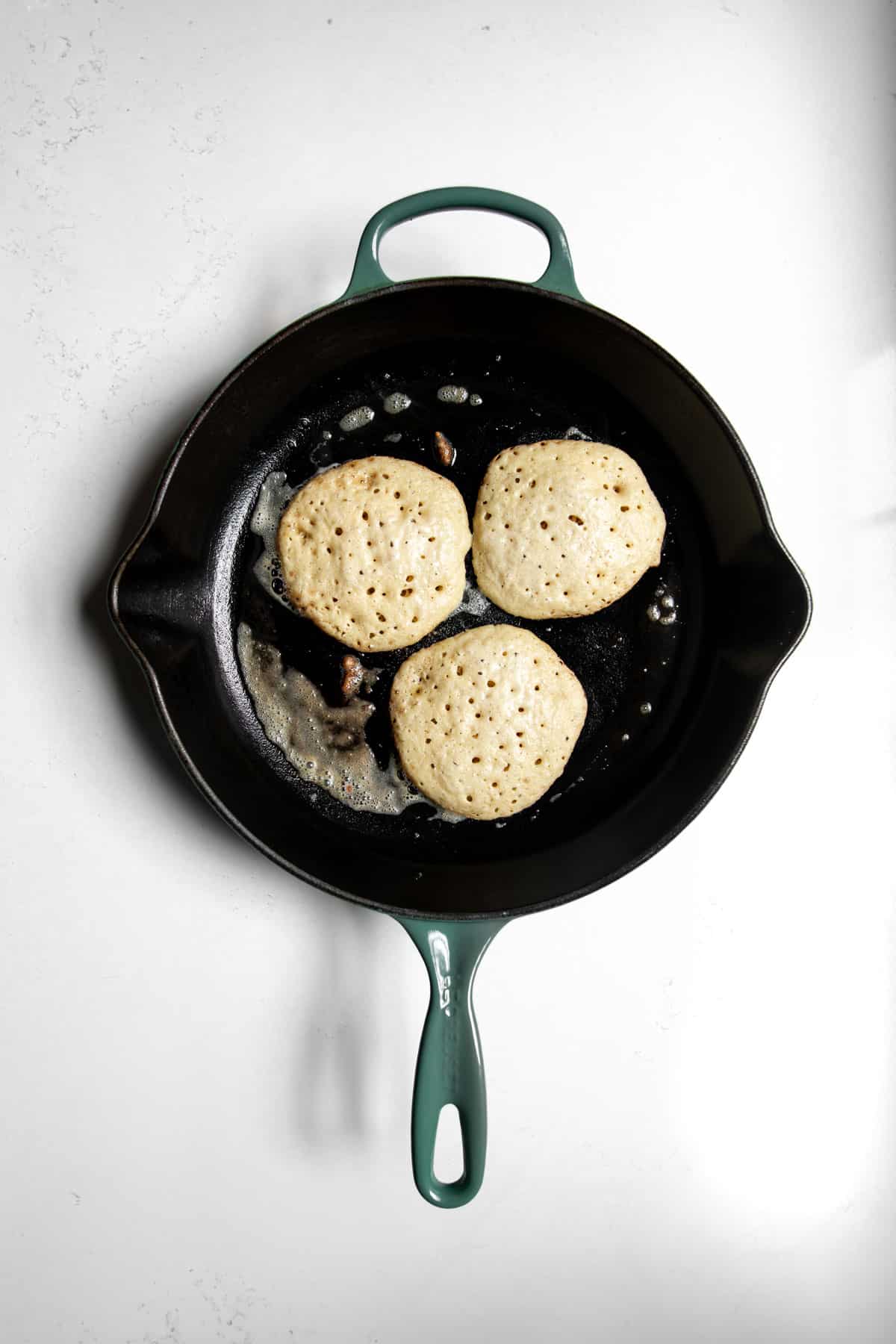 Three oat flour pancakes sitting on a skillet with bubbles before being flipped.