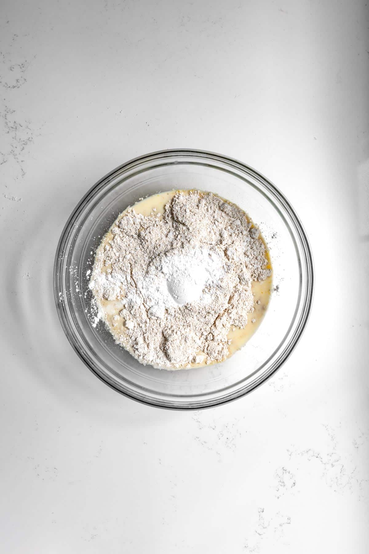 The wet ingredients of oat flour pancakes topped with dry ingredients of oat flour pancakes sitting in clear bowl on marble counter.