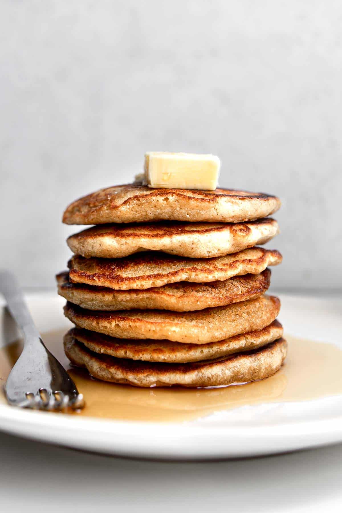 Close up of stack of seven oat flour pancakes sitting on a beige plate with a fork topped with a cube of butter and a drizzle of maple syrup.