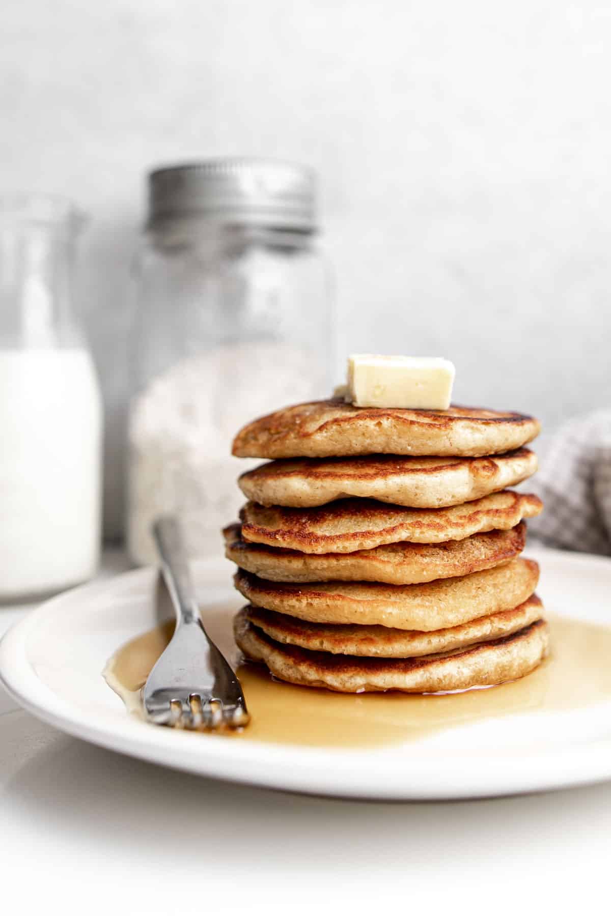 Stack of seven oat flour pancakes on a beige plate topped with butter and maple syrup with a jar of oat flour and a glass of milk in the background.