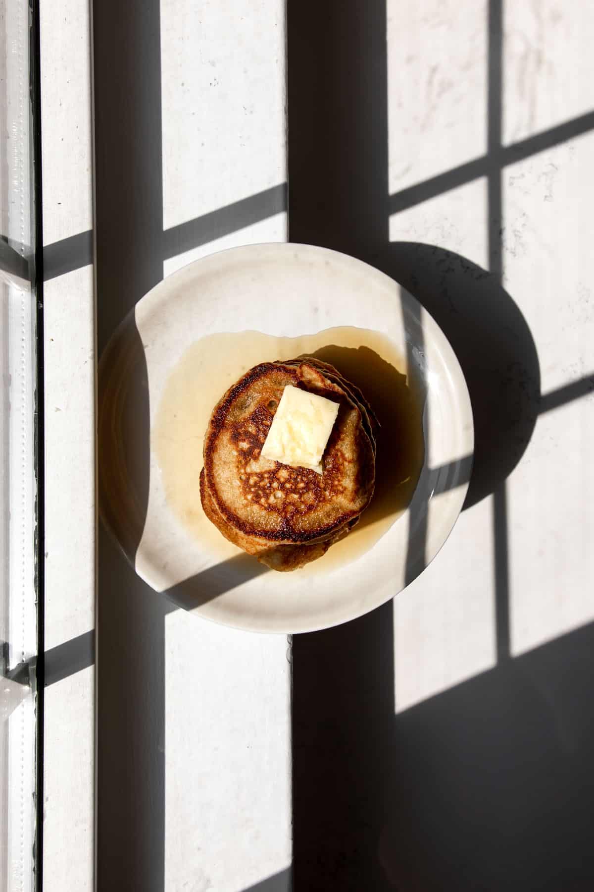 Overhead stack of oat flour pancakes sitting on a beige plate in front of a window with shadows casting.