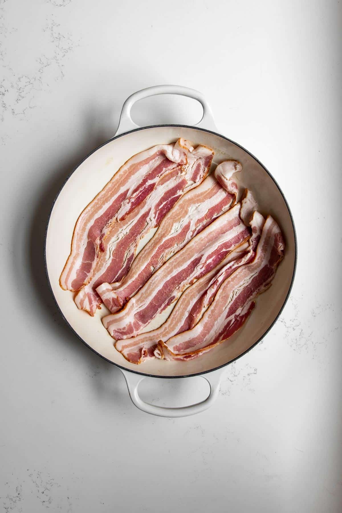 Strips of bacon in a pan on a white marble table cooking.