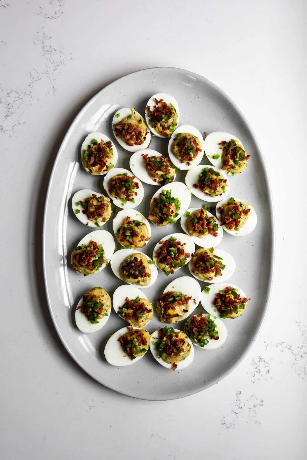 Devilled eggs topped with jalapeno and bacon on a grey platter on a marble table.