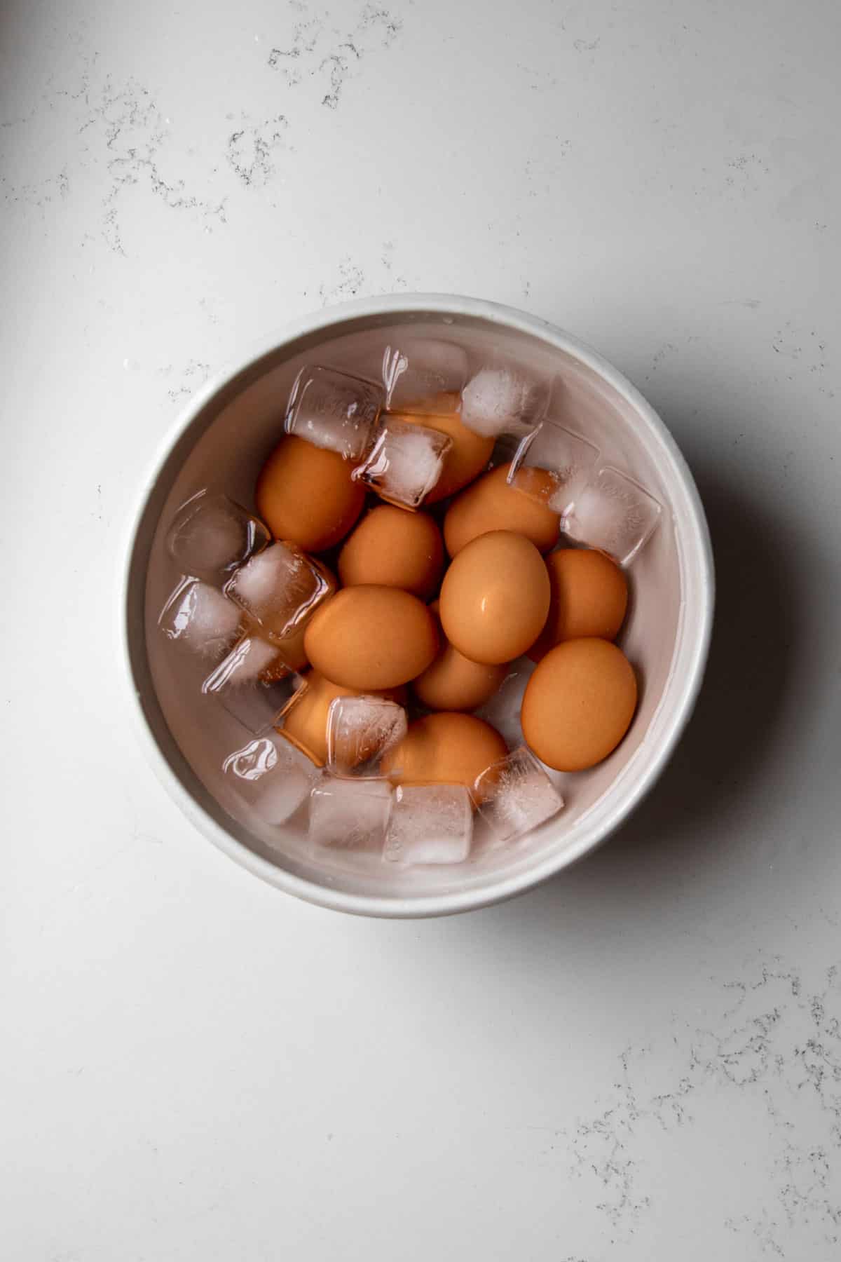 Boiled eggs in a white bowl filled with cold water and ice on a marble table.