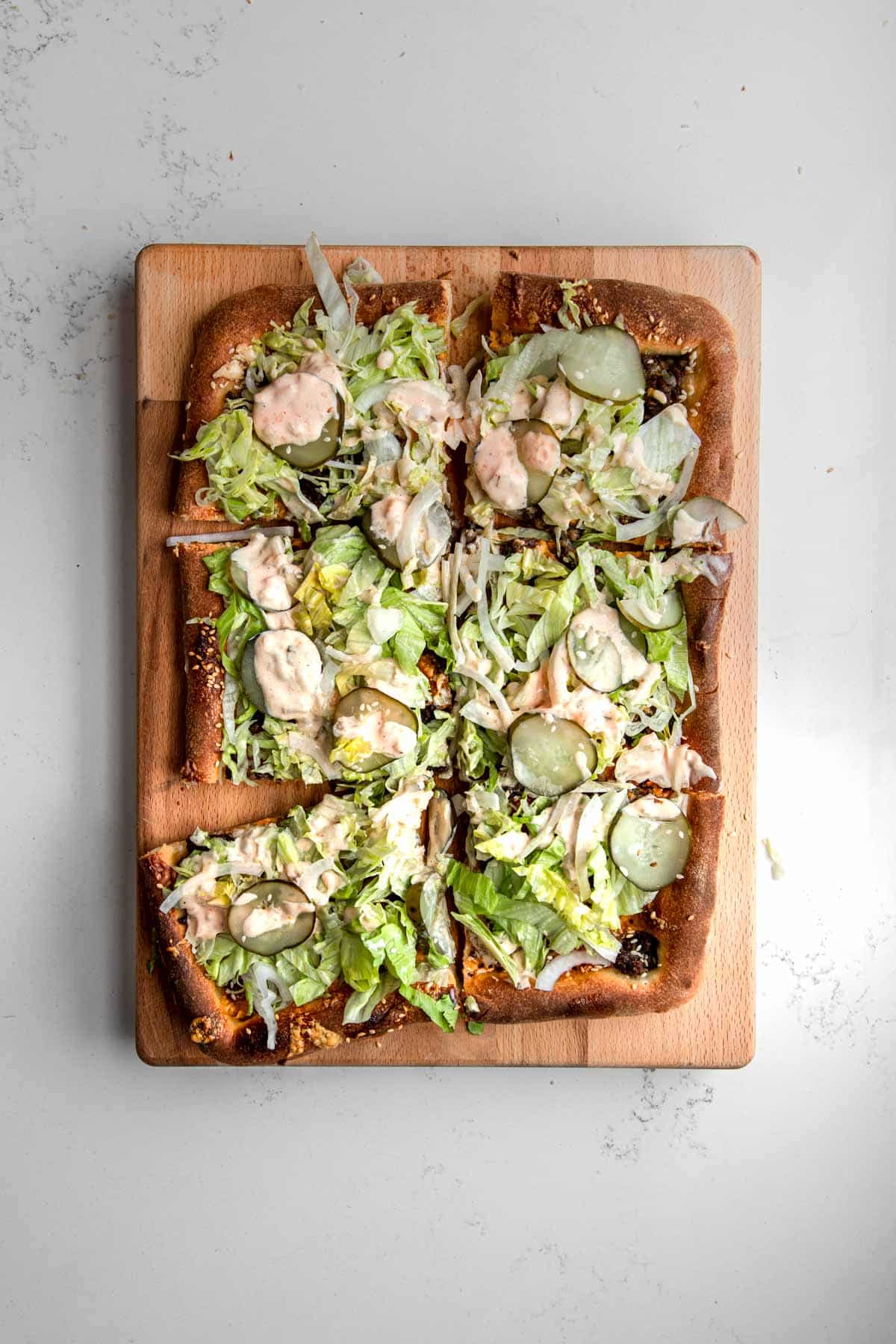 Big Mac Pizza cut into six squares on a wooden cutting board topped with lettuce, pickles, onions and big mac sauce.