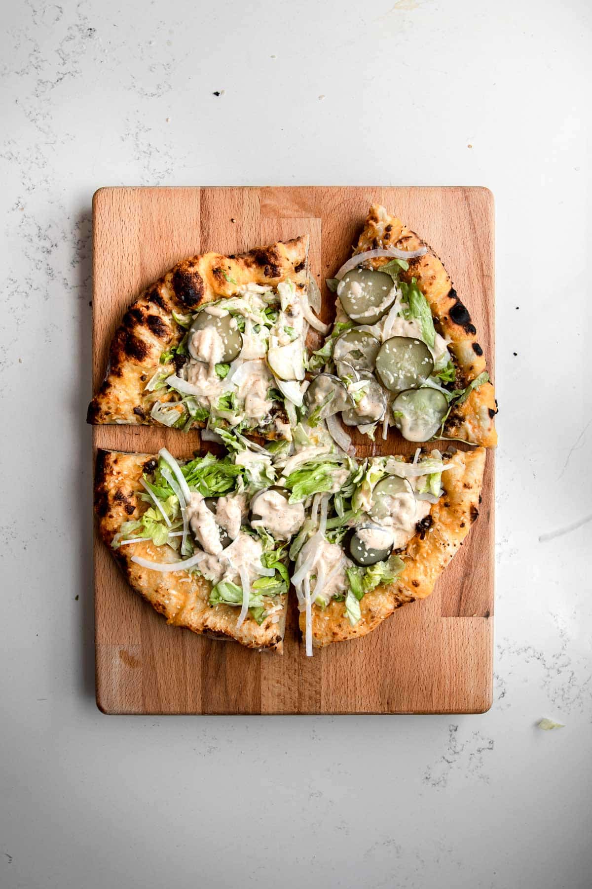 A Neapolitan-style big mac pizza cut into 4 slices with wood fired crust on a white plate topped with lettuce, pickles, onions and big mac sauce.