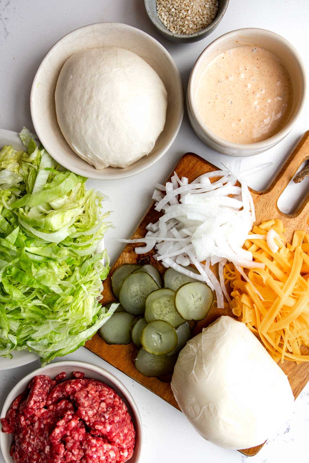 Ingredients needed to make a Big Mac Pizza on a table. Pizza dough, Big Mac sauce, lettuce, ground beef, onions, pickles, grated cheddar cheese and mozzarella cheese ball. 