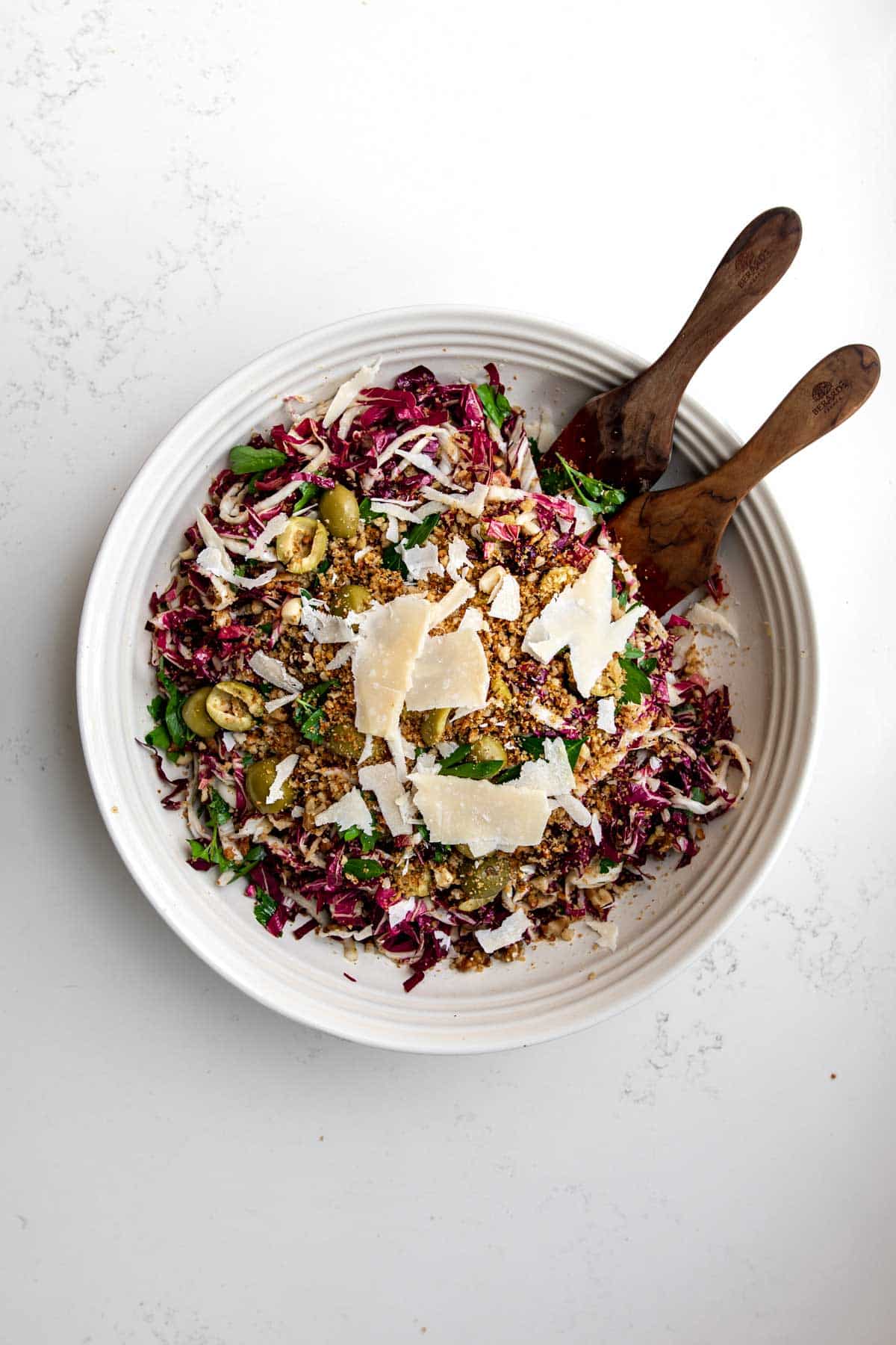 Large white bowl filled with radicchio salad topped with parmesan, olives and breadcrumbs with two wooden spoons coming out of the side.