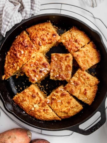 Sweet Potato Cornbread cut in squares in a cast-iron skillet on a wire rack with a gingham tea towel wrapped around the handle.