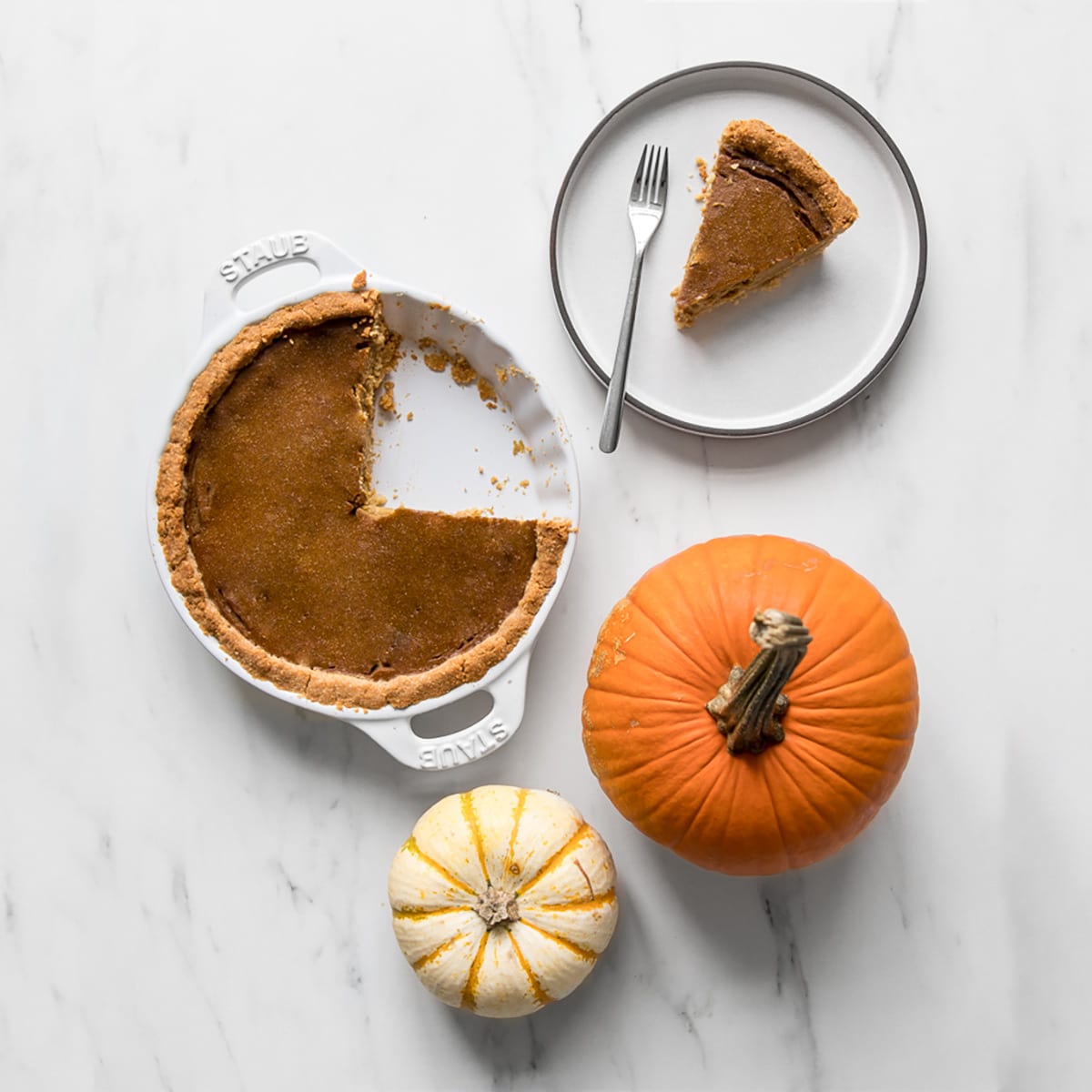 Pumpkin pie in a white pie dish, with slice removed and on a grey plate. Two small pumpkins placed beside the pie dish.