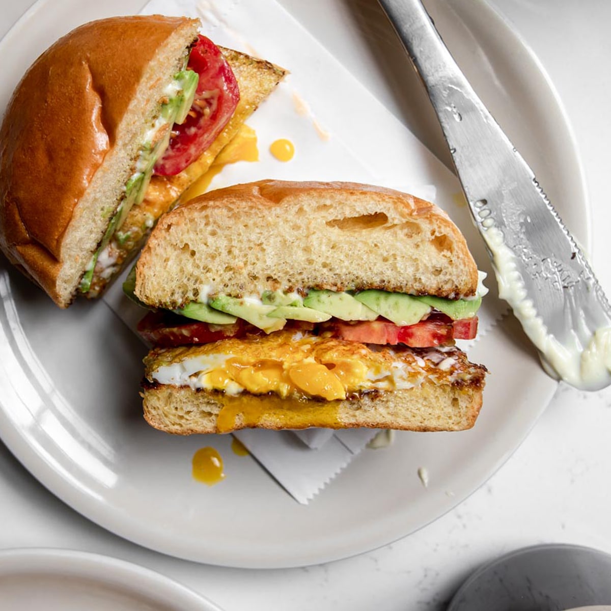 cut open fried egg sandwich with mayo, avocado, and tomato on a plate.