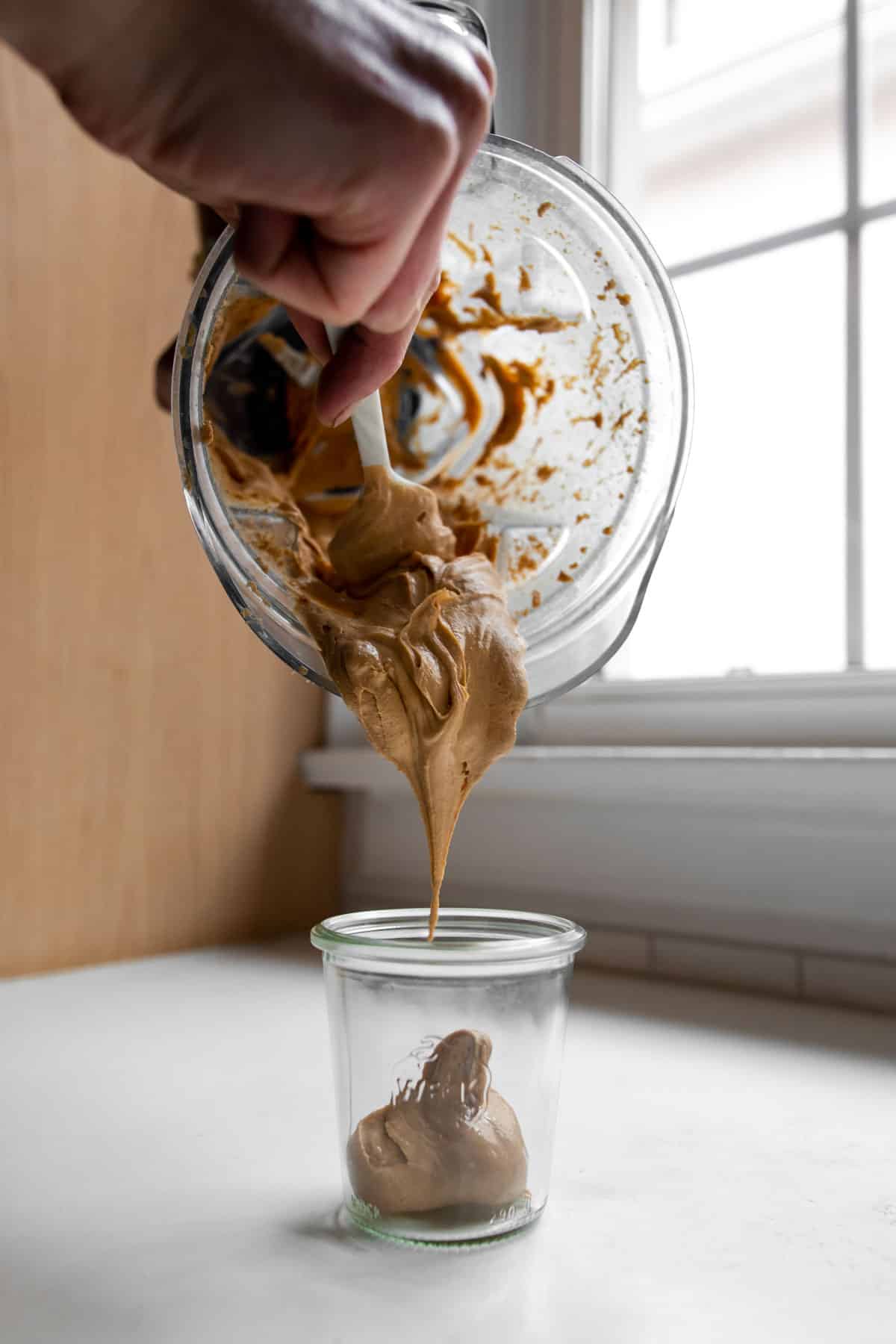 Transferring cashew butter from a blender container to a glass jar in front of a window.