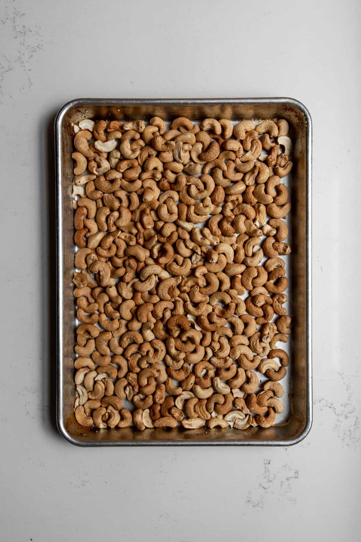 Overhead view of roasted cashews on a silver baking sheet.