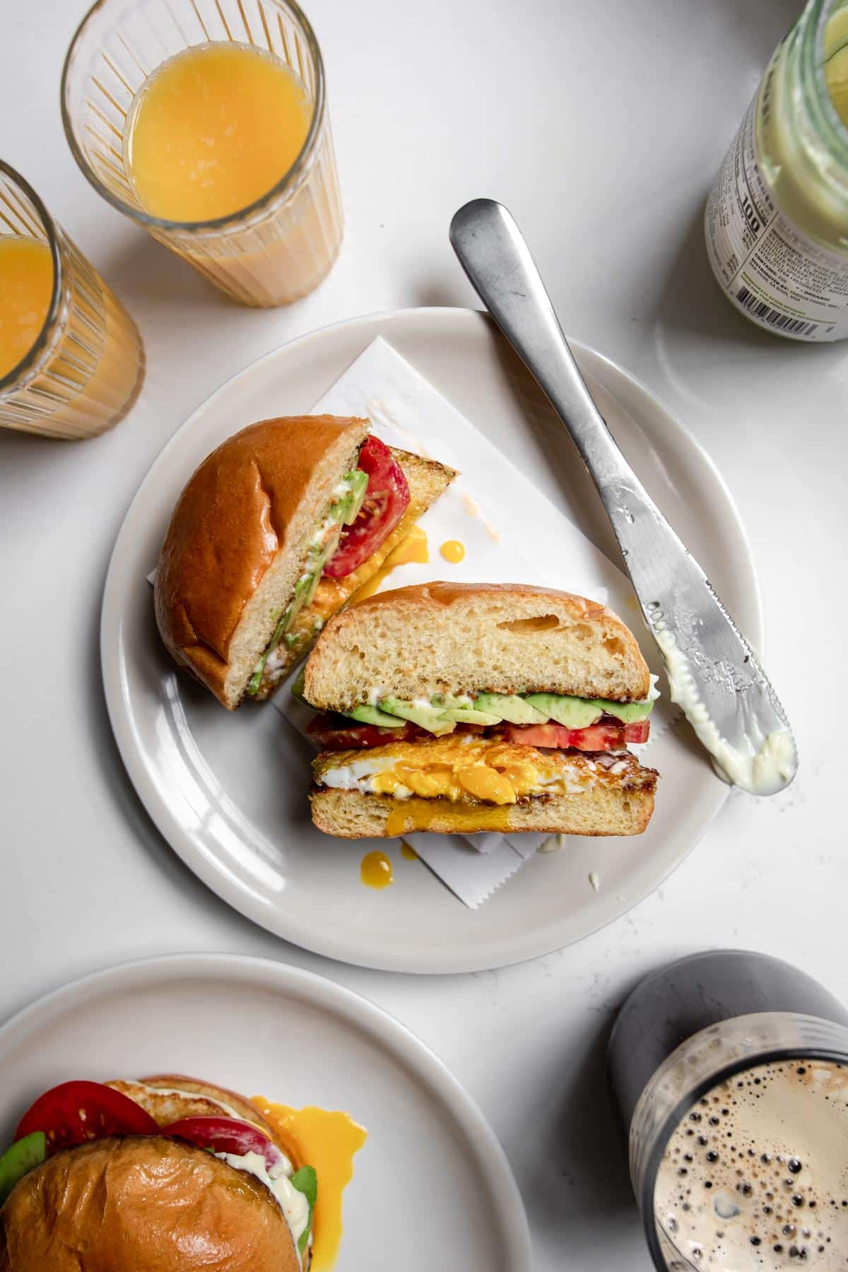 Breakfast sandwich cut in a half with avocado, tomato and fried egg on a white plate.