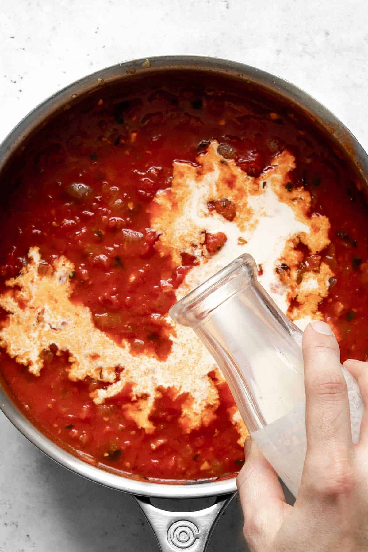 hand adding cream to tomato sauce in pan.