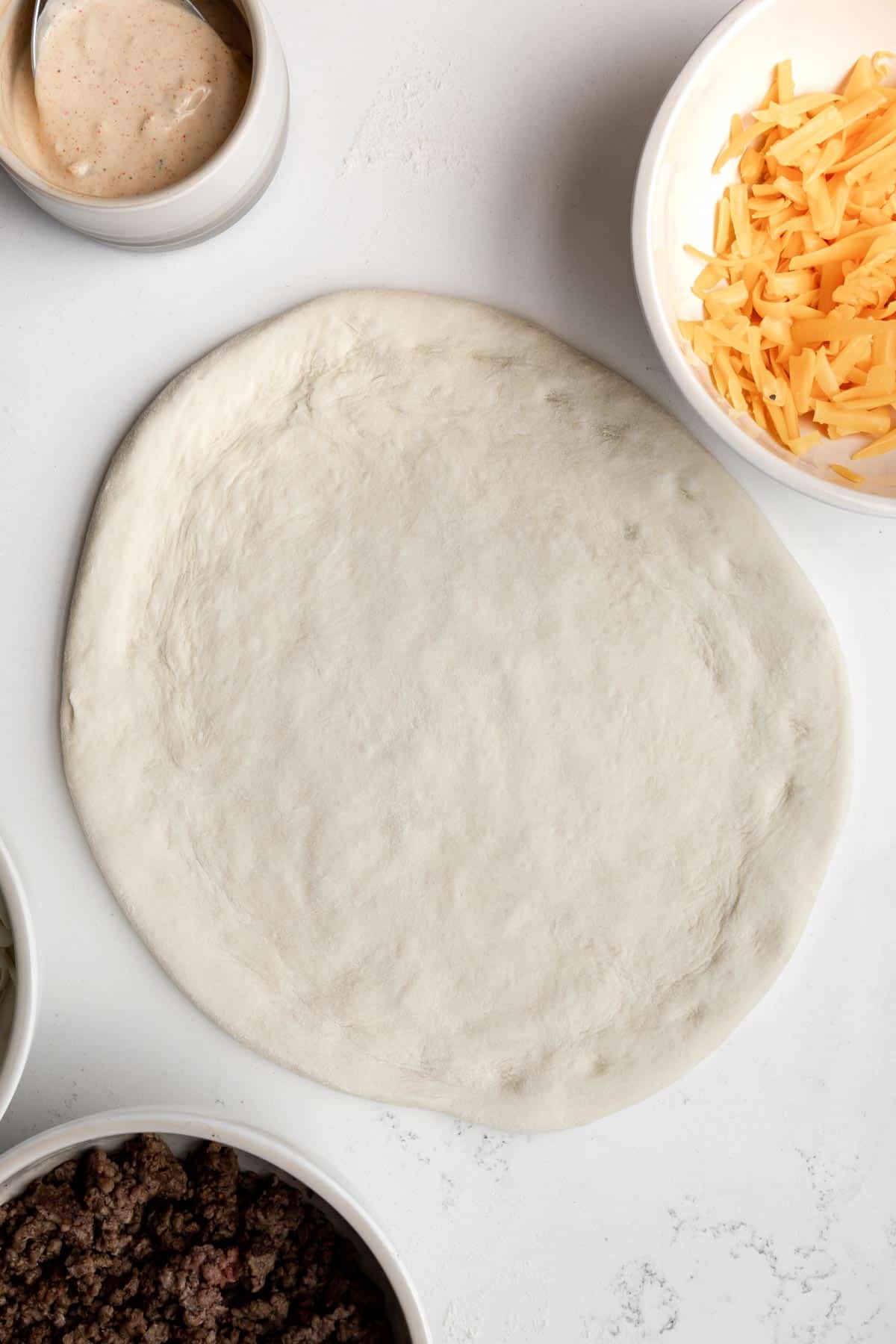 pizza dough stretched on a table with ingredients around it.