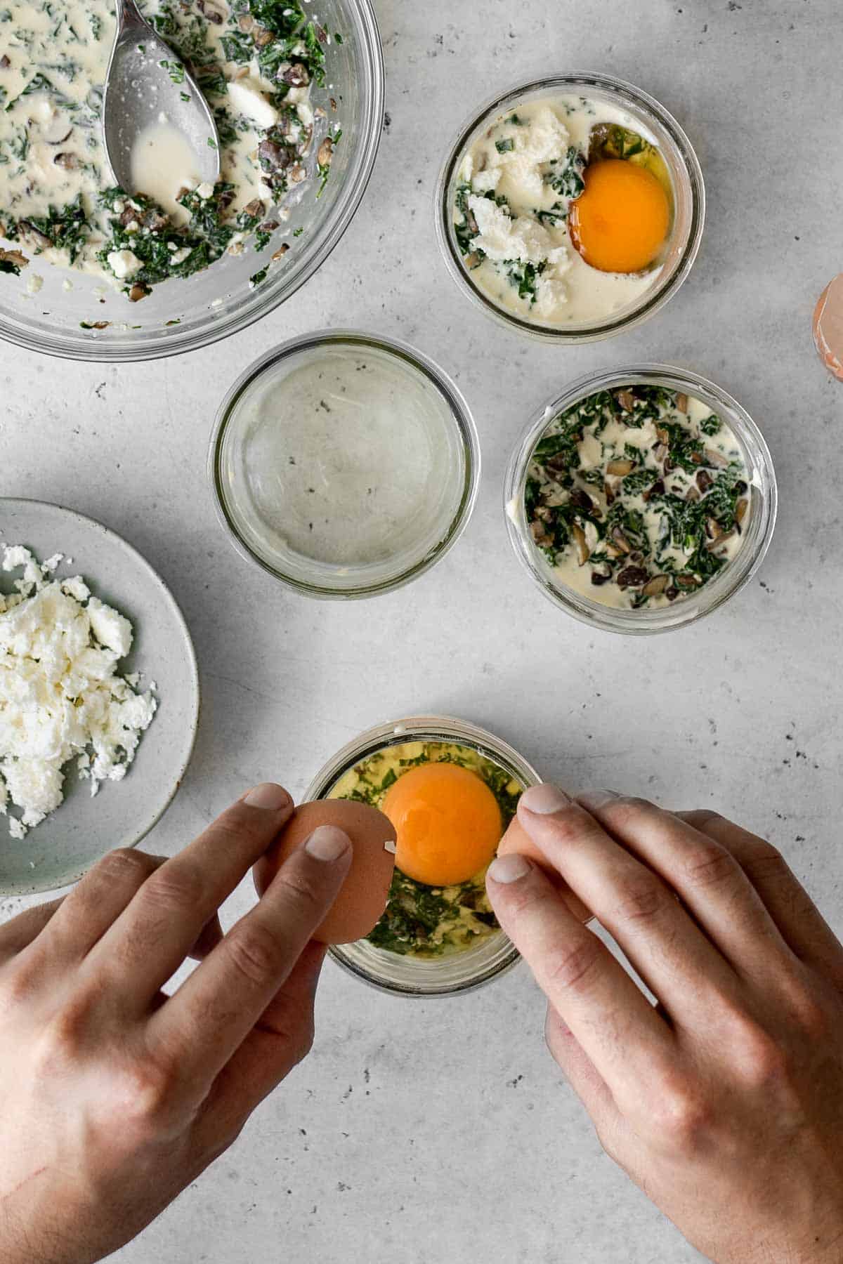 An egg being cracked into a mason jar with cream, mushroom, kale and feta.