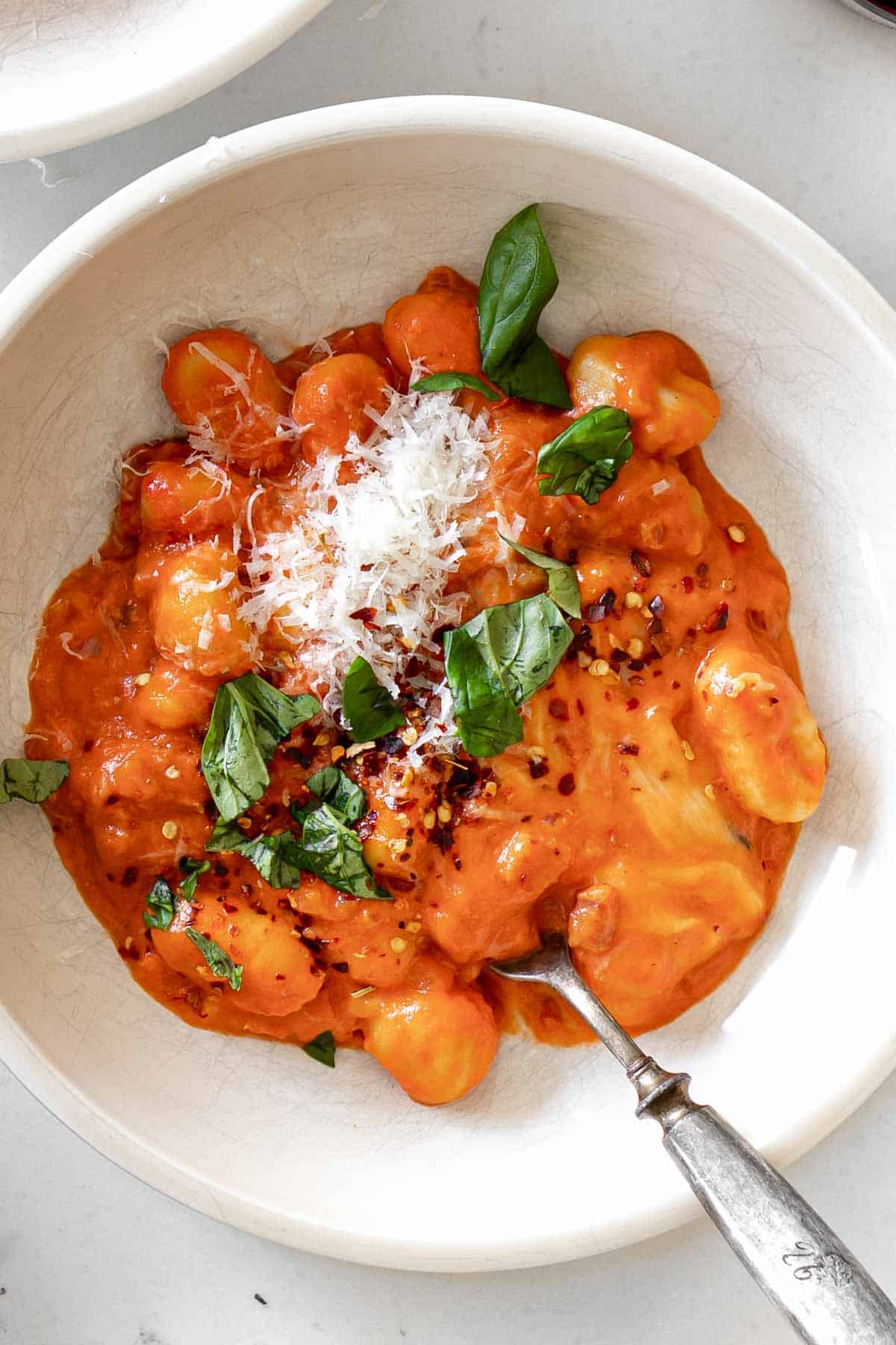 close up gnocchi in a pasta bowl with a fork topped with basil, parmesan and red pepper flakes.