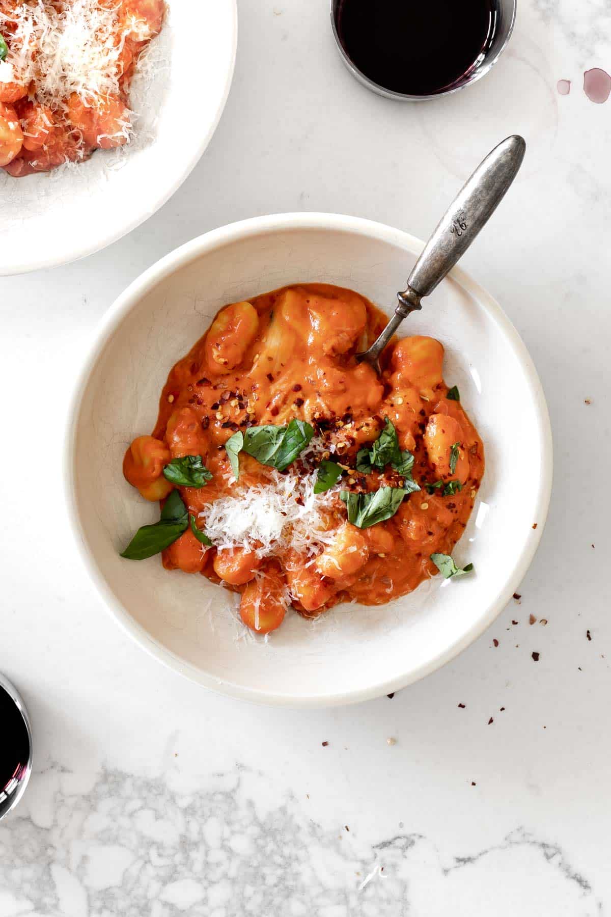 gnocchi in a pasta bowl with a fork topped with basil, parmesan and red pepper flakes with a glass of wine next to it.