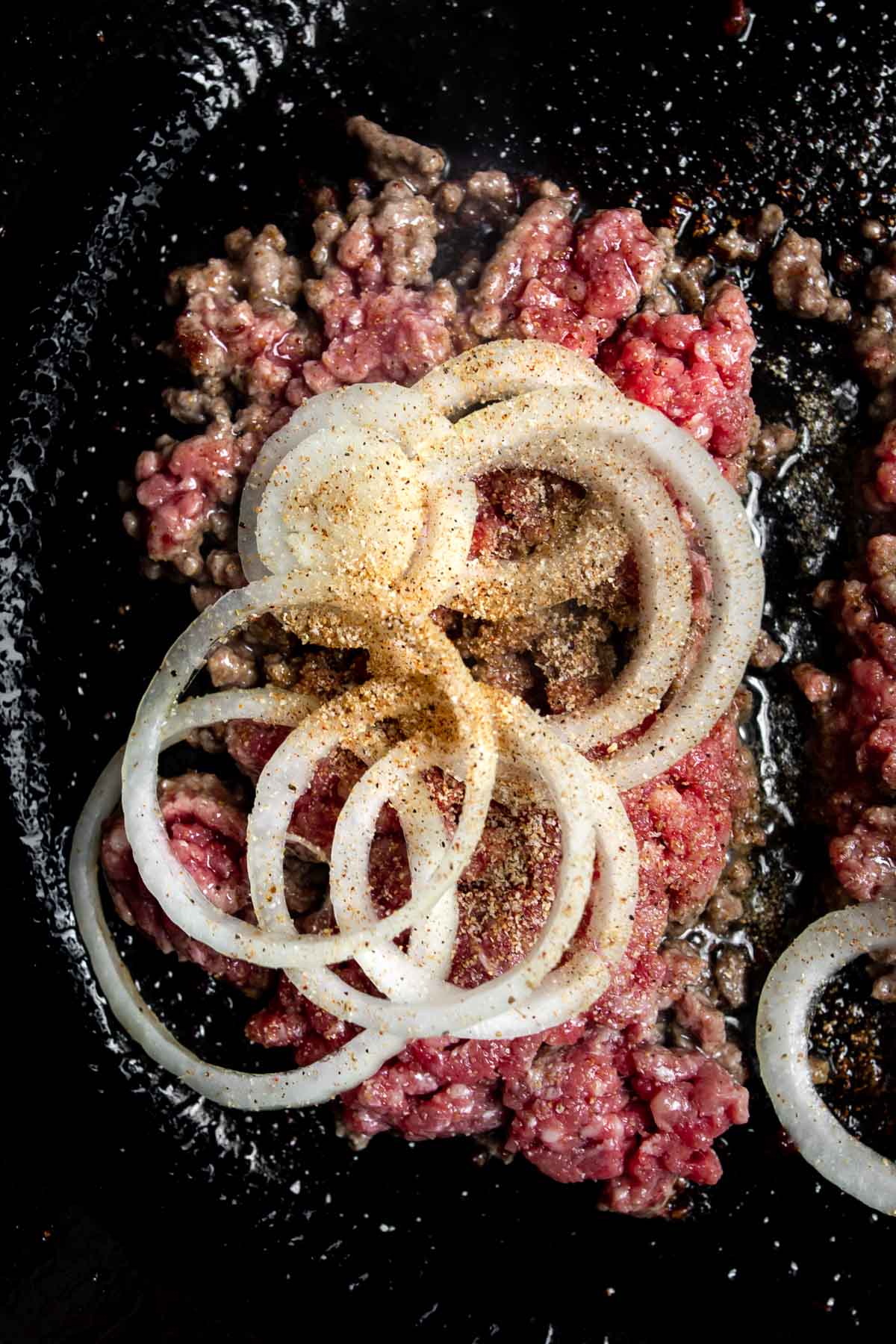 Onions topped with adobo seasoning on ground beef cooking in a cast iron skillet.