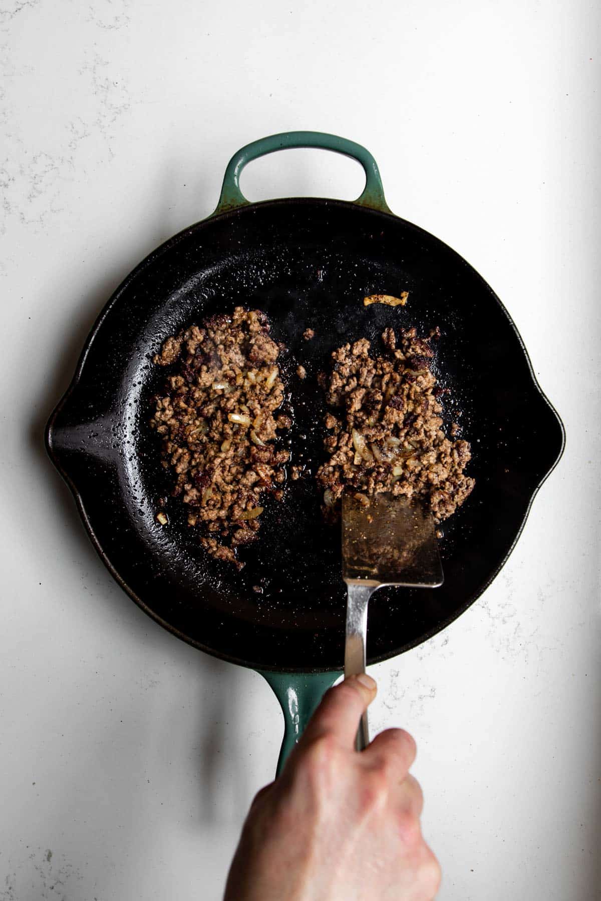 Metal spatula chopping the ground beef and onion in a cast iron skillet.