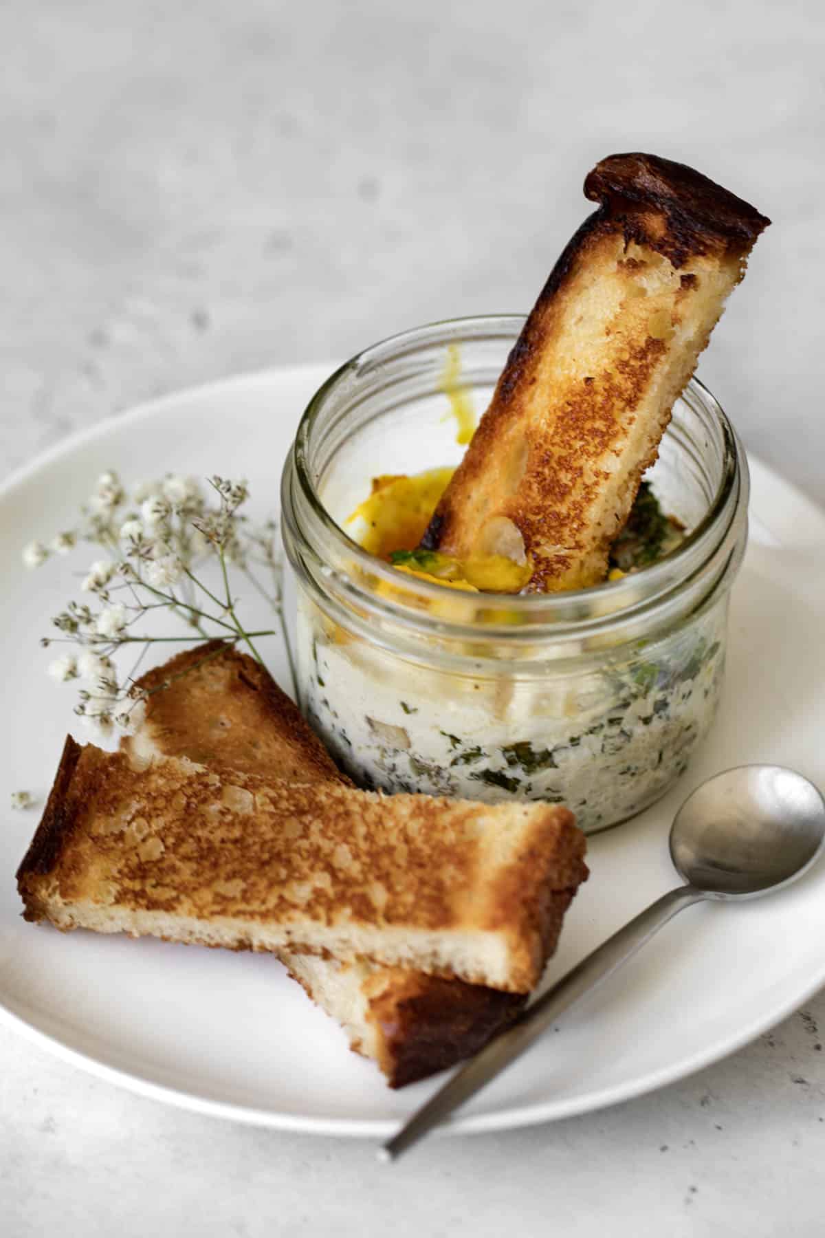 coddled eggs with mushroom, kale and feta in a mason jar on a plate with a piece of toast breaking open the yolk.