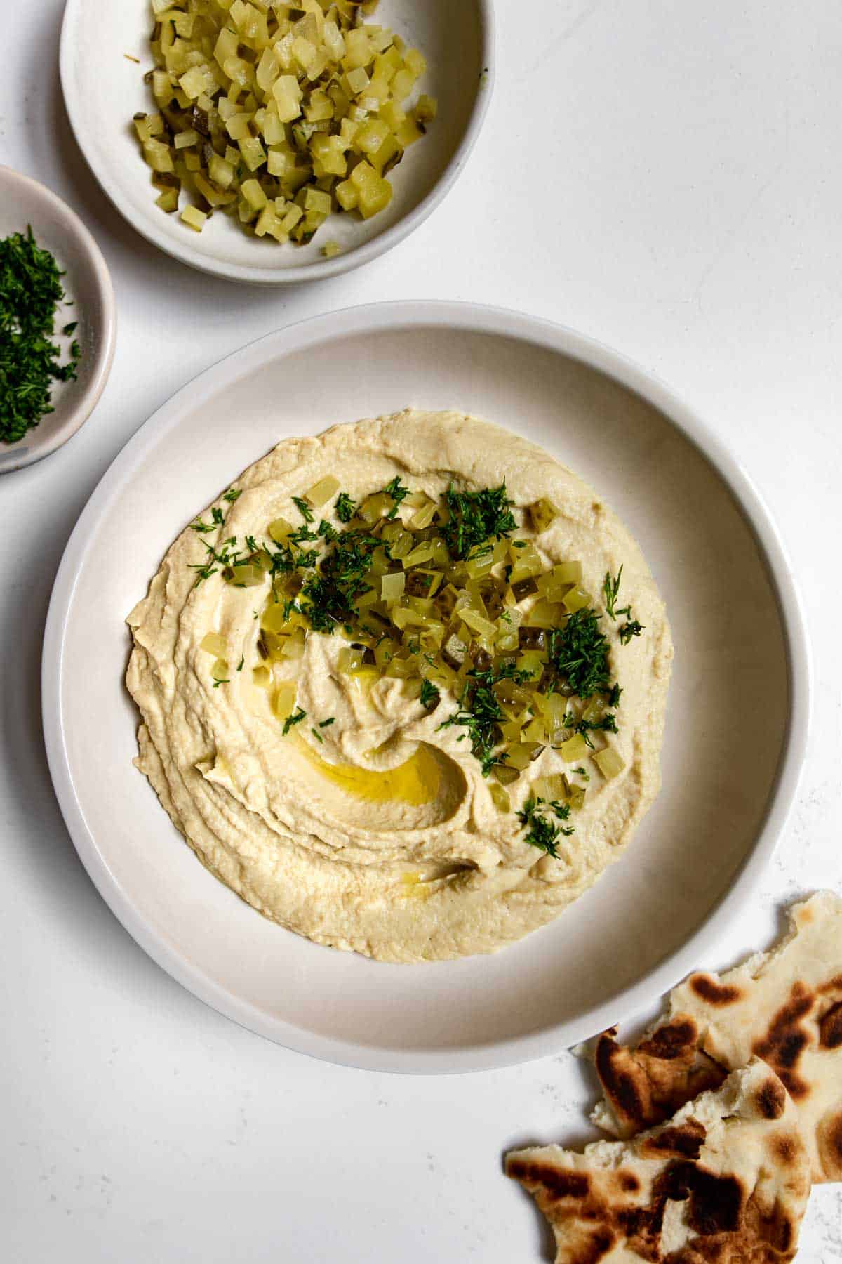 hummus topped with chopped dill pickle and dill sprigs with pita bread next to it.