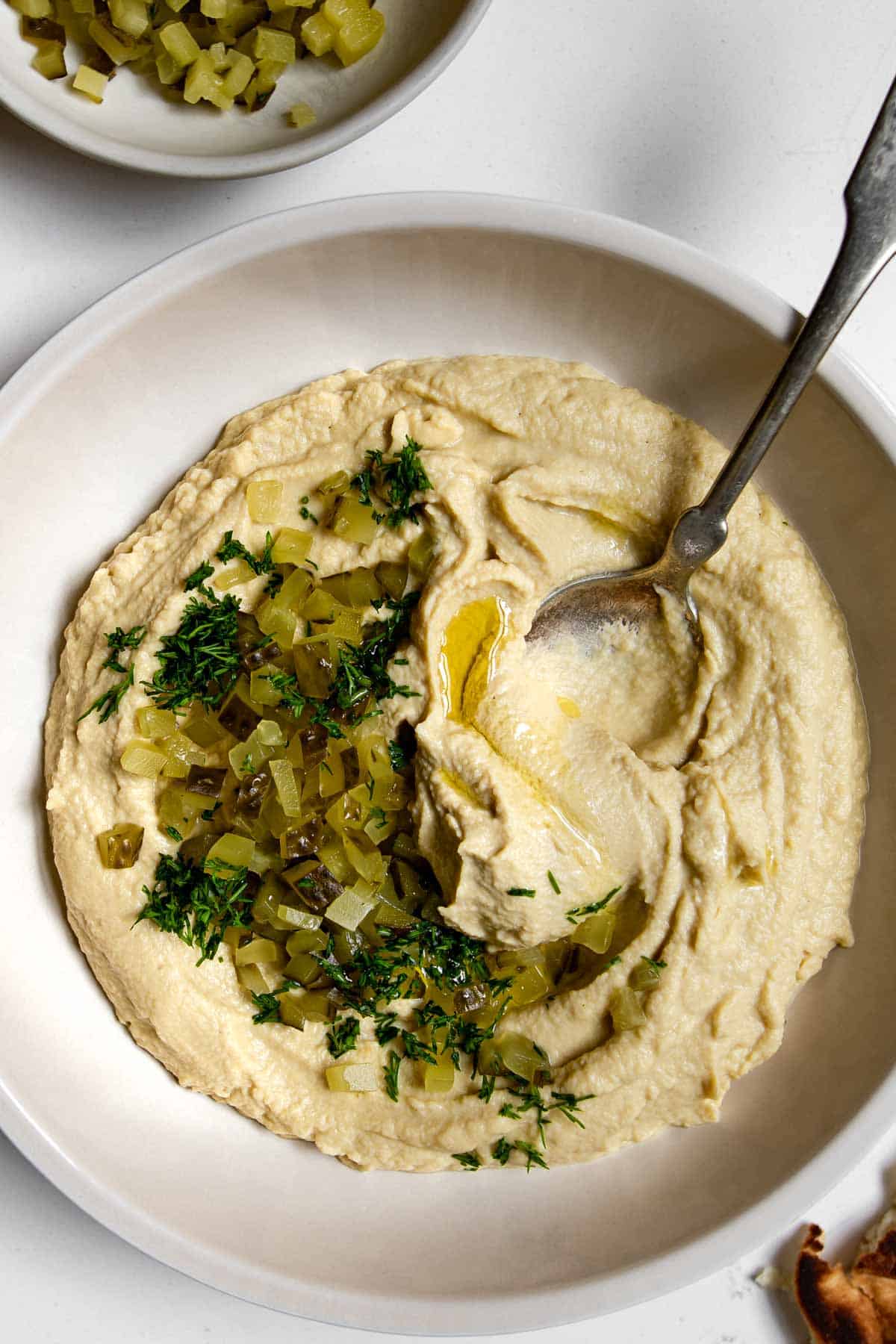 close up of bowl filled with hummus topped with dill pickles and dill with a spoon in it.