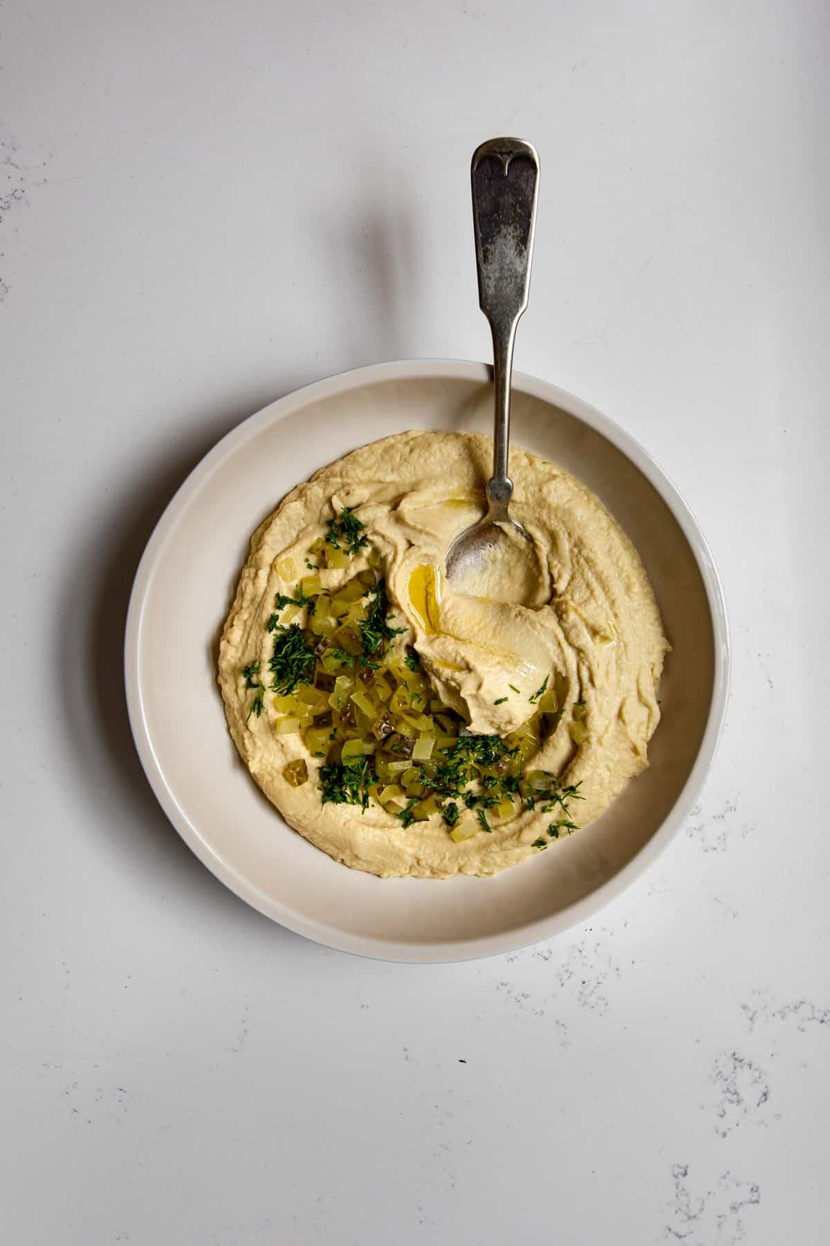creamy hummus on a plate topped with dill pickles and dill with a spoon.