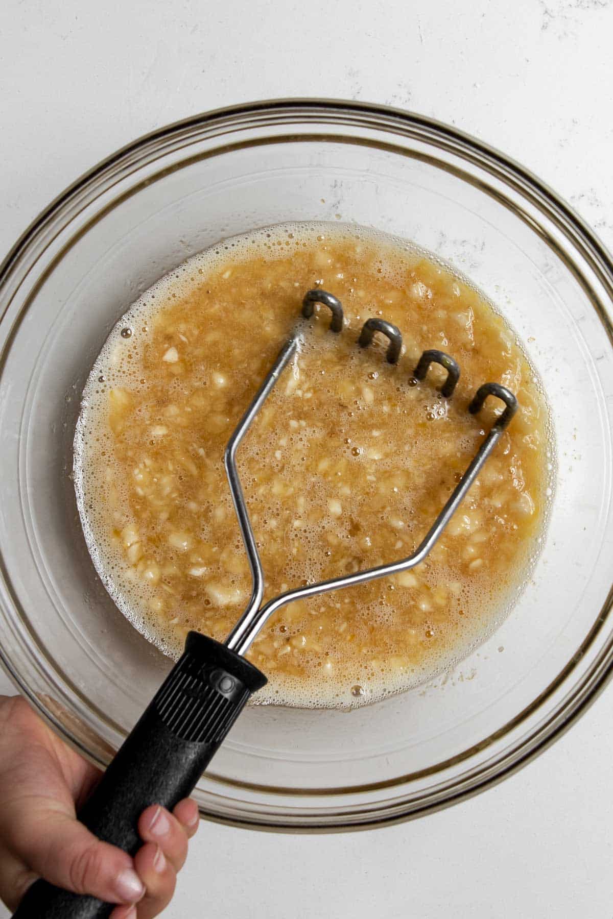 child mashing wet ingredients of bananas, egg, and maple syrup in a bowl.