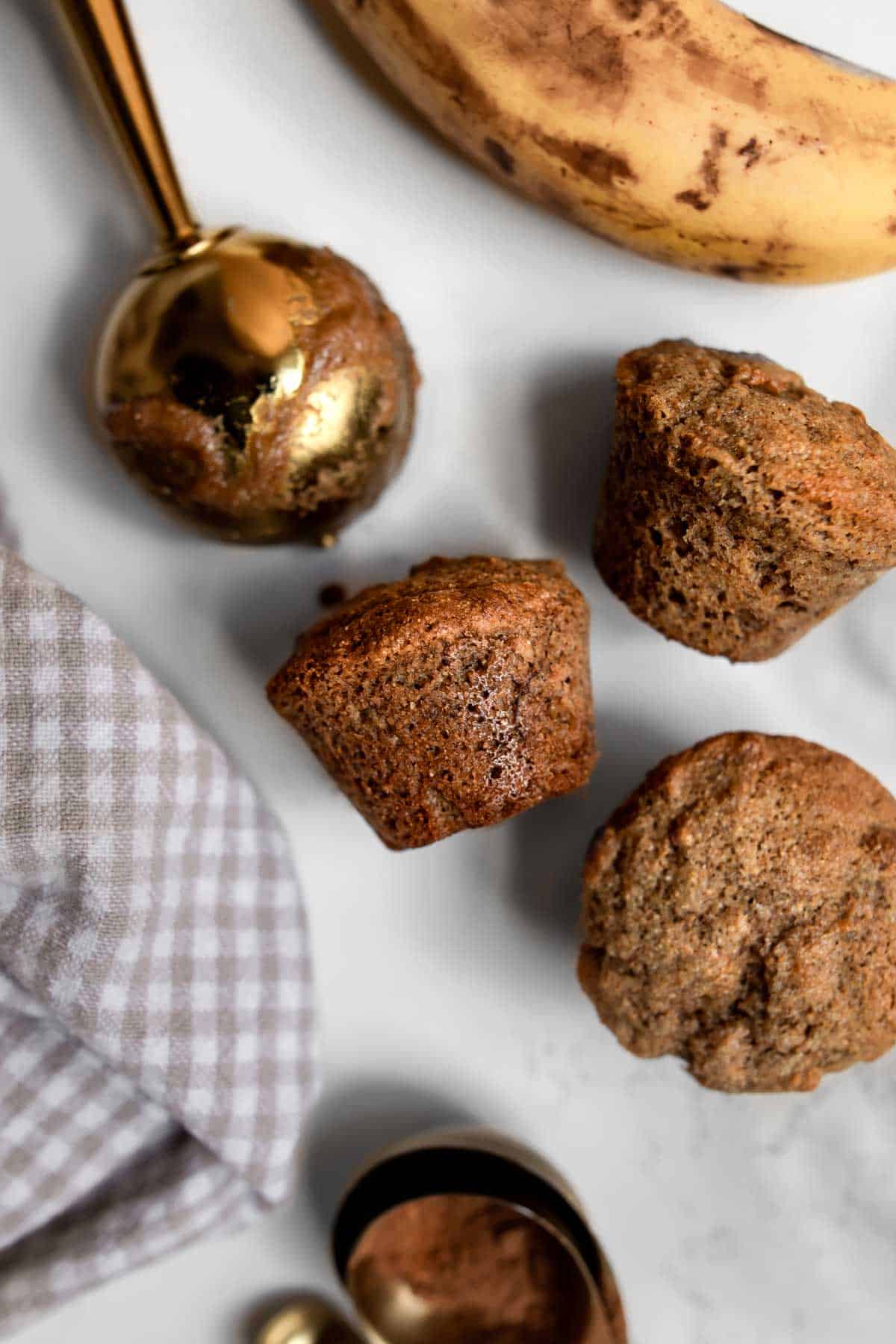 mini banana muffins on a table with a scoop, banana and a cloth around it.