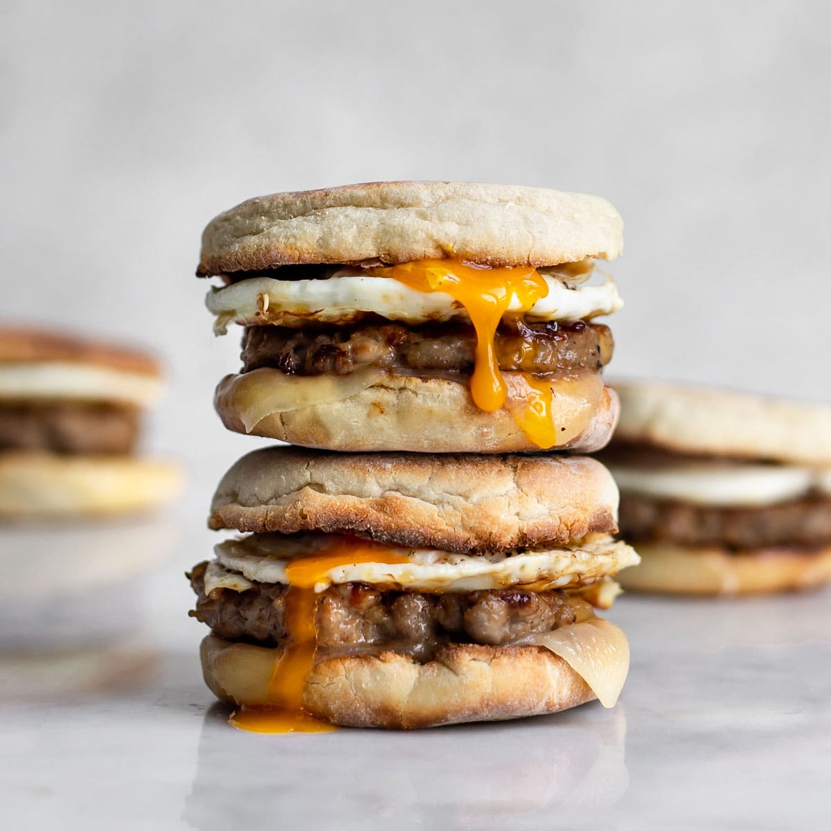 Two sausage, egg and cheese breakfast sandwich on an english muffin stacked on top of each other with yolk running down.