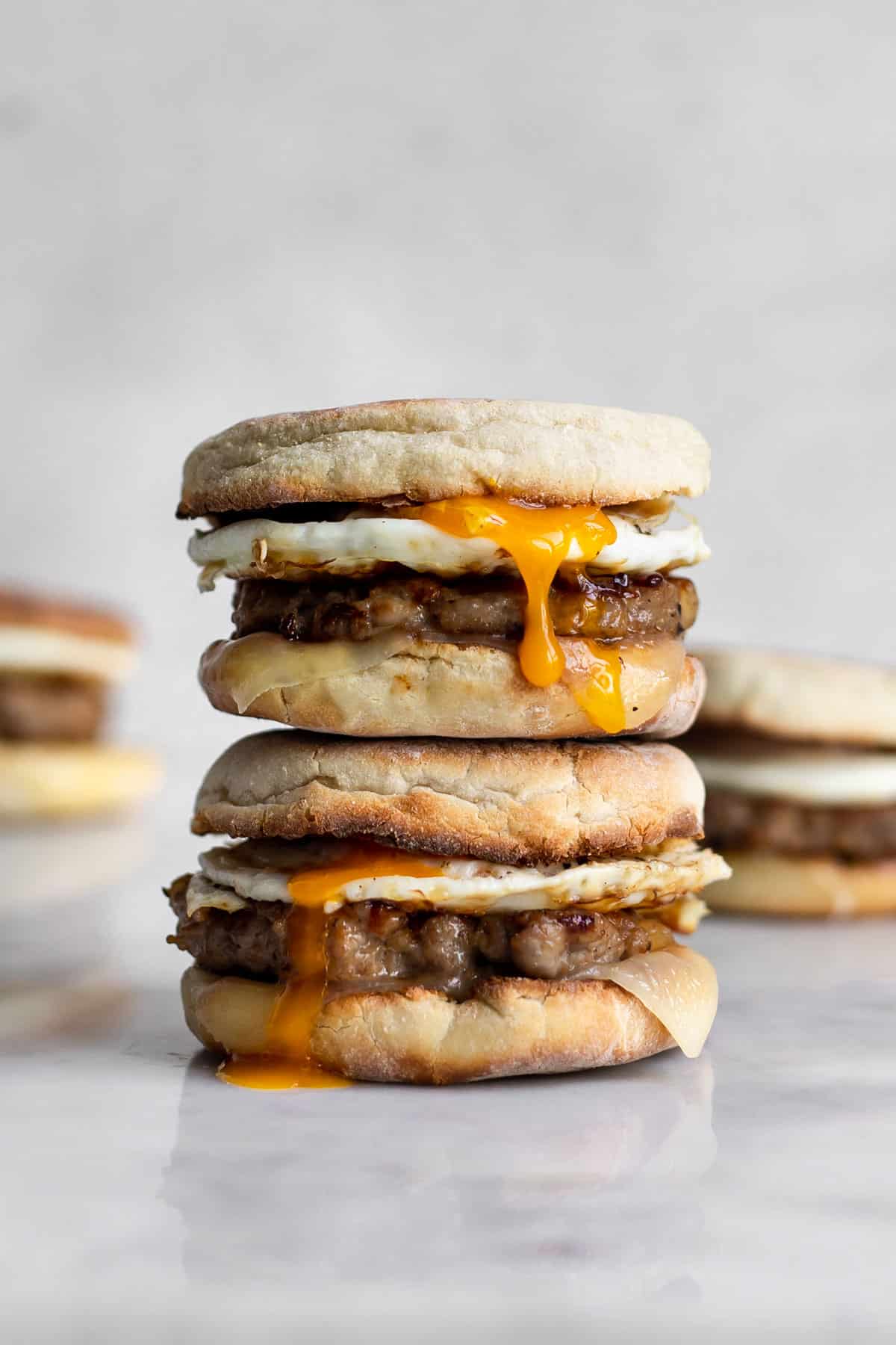 Two sausage, egg and cheese breakfast sandwich on an english muffin stacked on top of each other with yolk running down.