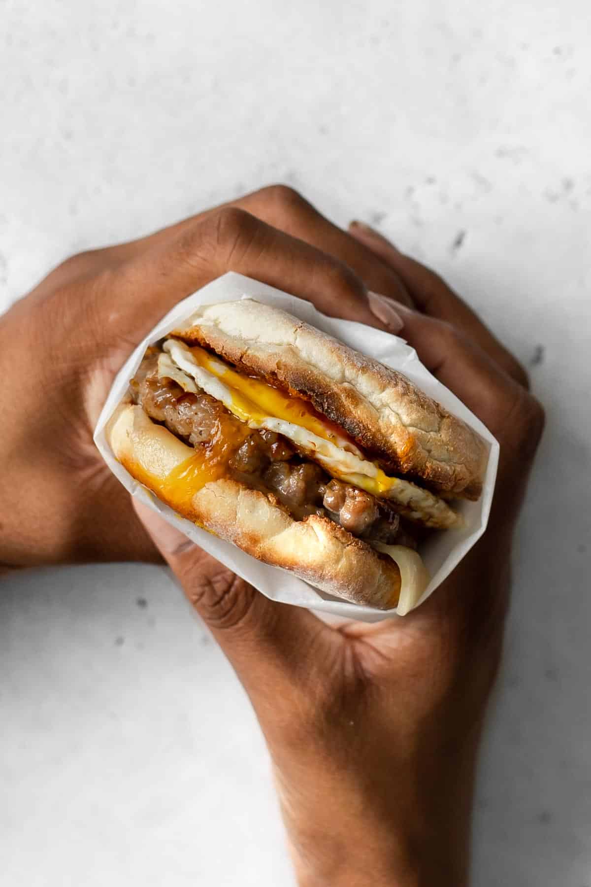 Hands holding a sausage, egg, and cheese mcmuffin breakfast sandwich wrapped in parchment.