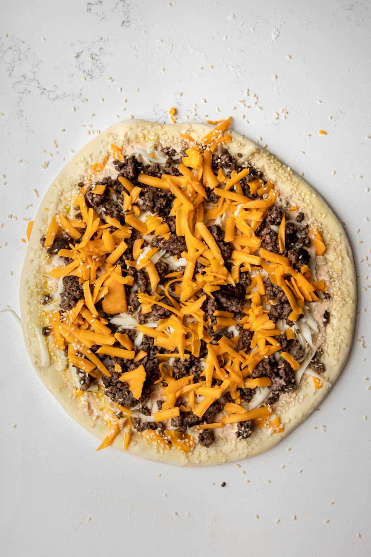 pizza dough topped with big mac sauce, mozzarella cheese, cooked ground beef, cheddar cheese and sesame seeds.