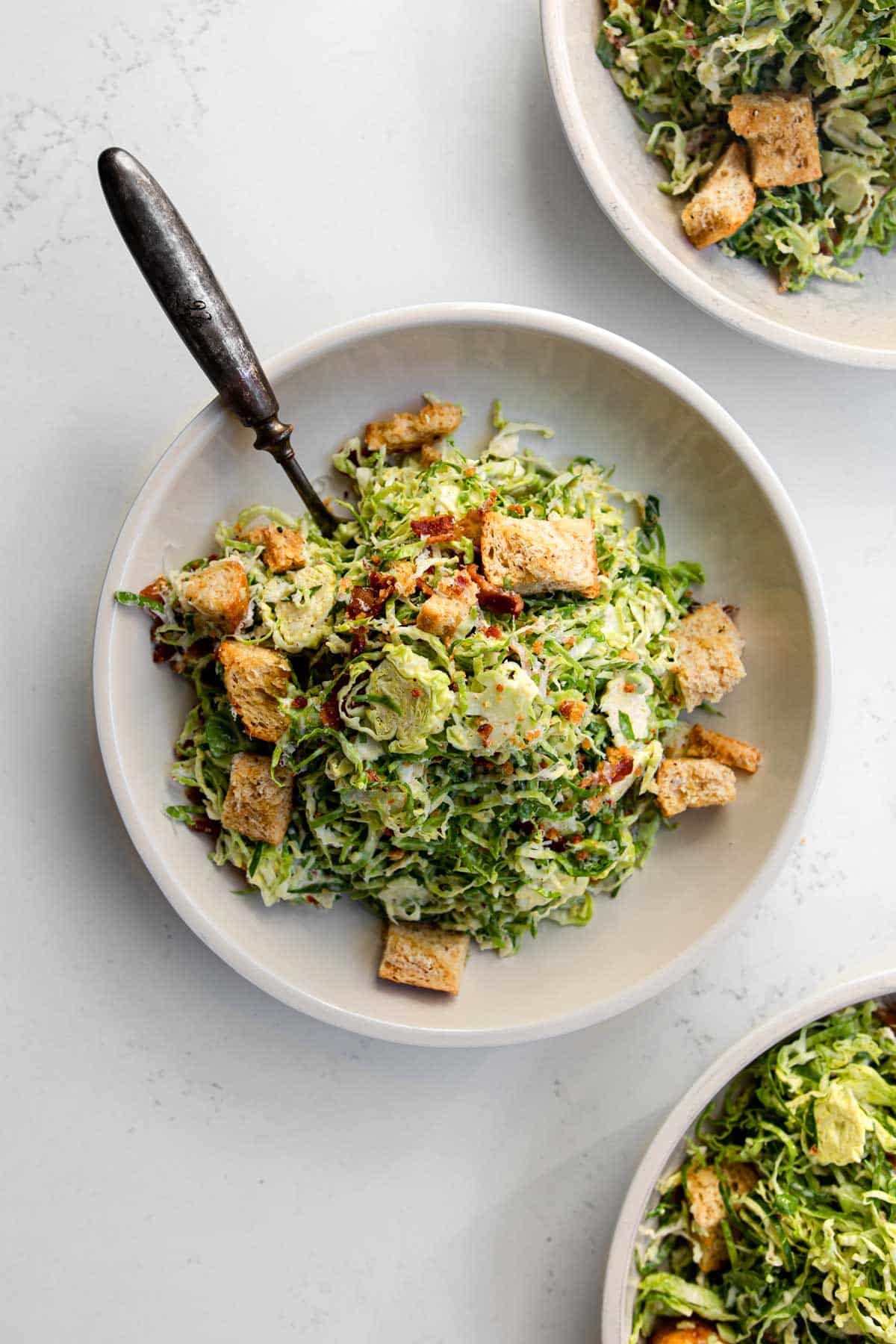 three bowls of brussels sprout caesar salad in plates with a fork.