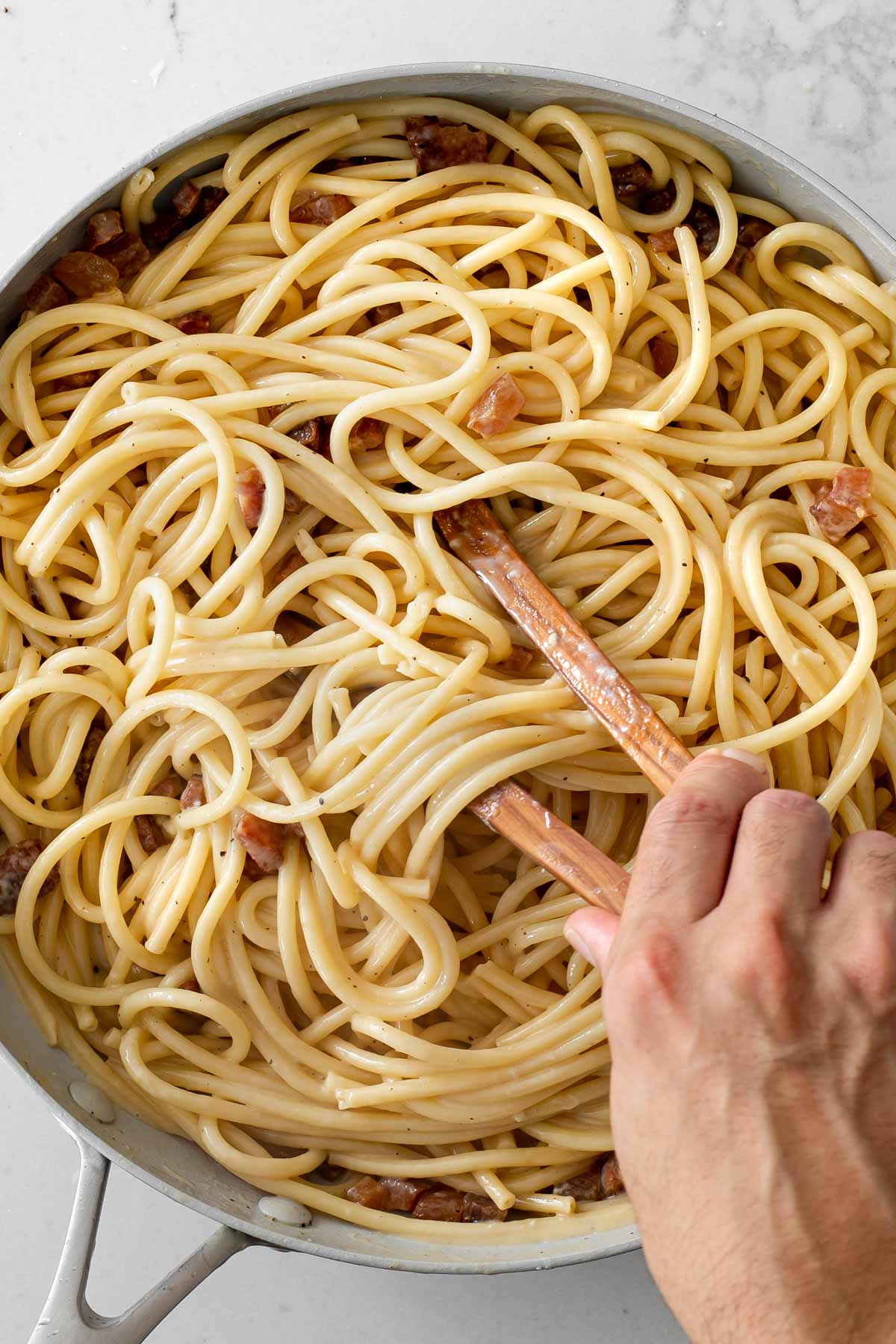 hand tossing egg mixture with guanciale and noodles in a pan using tongs.