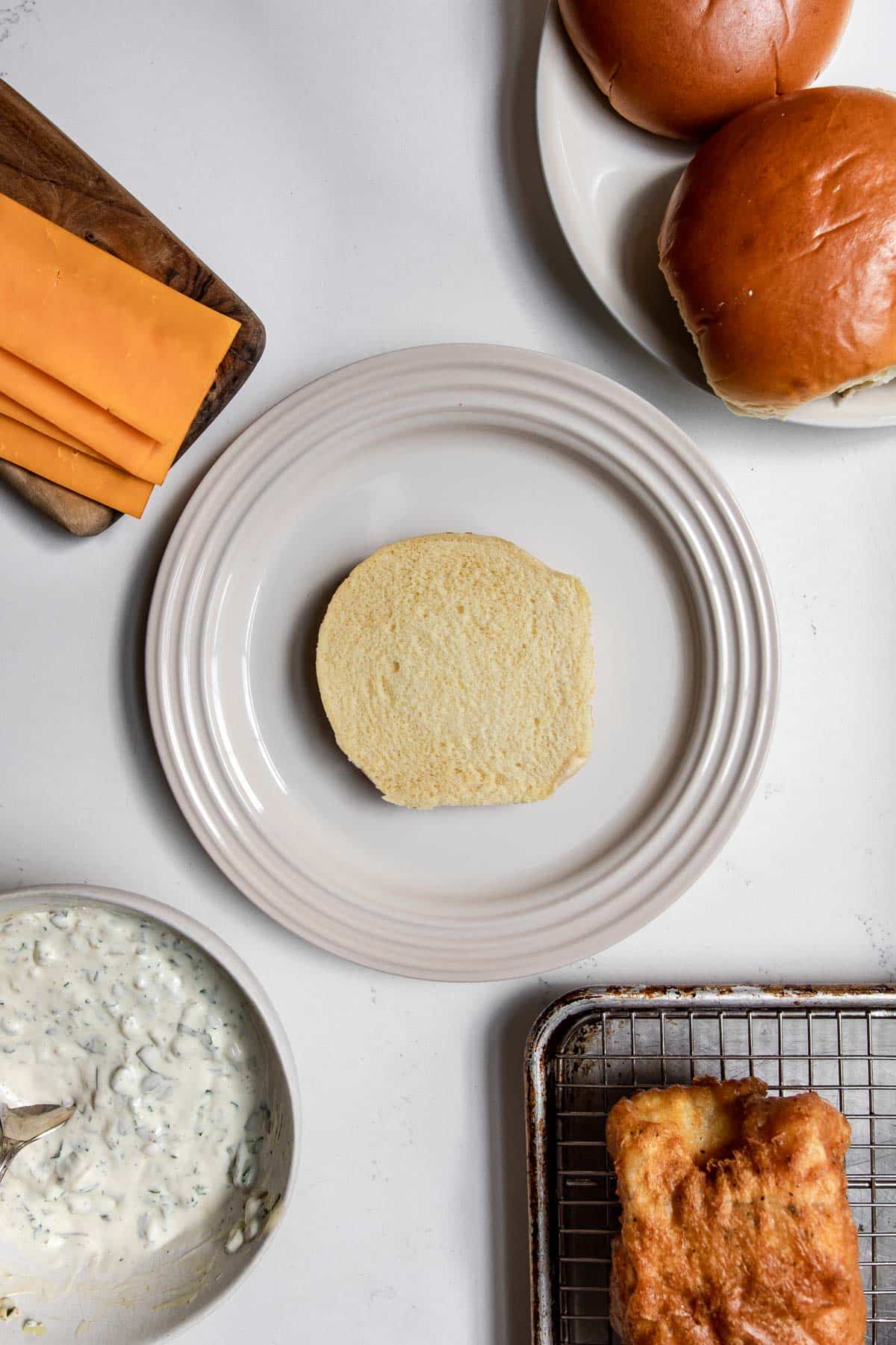 brioche bun on a plate with cheese, tartar sauce and fried cod around it.