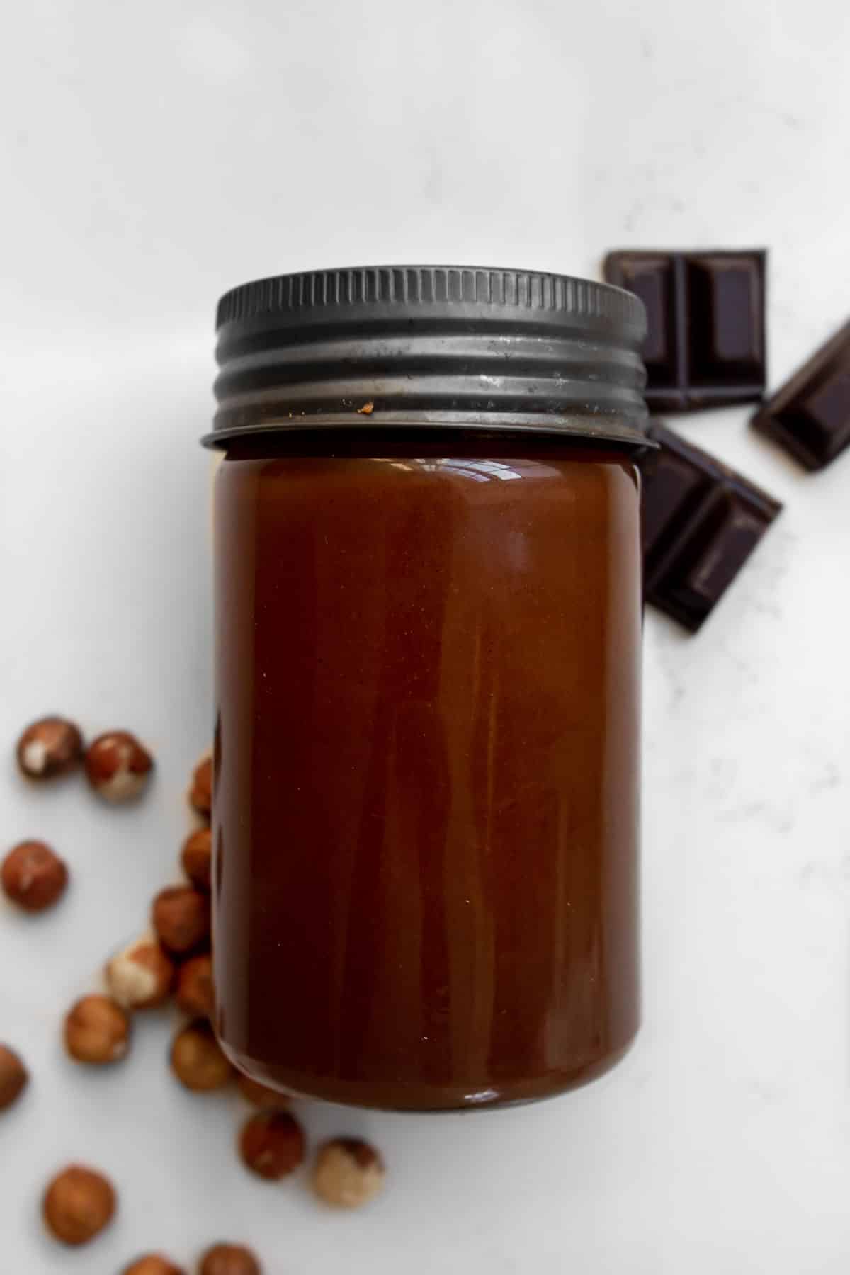 Jar of homemade chocolate hazelnut spread on a counter with hazelnuts and chocolate around it.