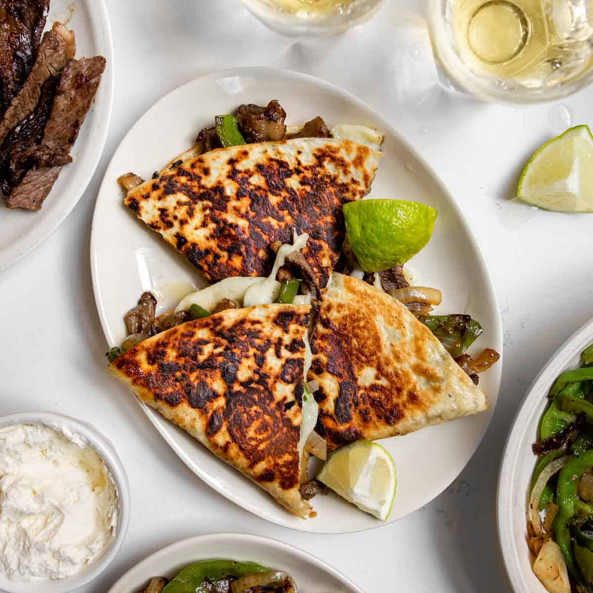Three Philly Cheesesteak Quesadillas cut and arranged on a plate with lime wedges, beer and ingredients.